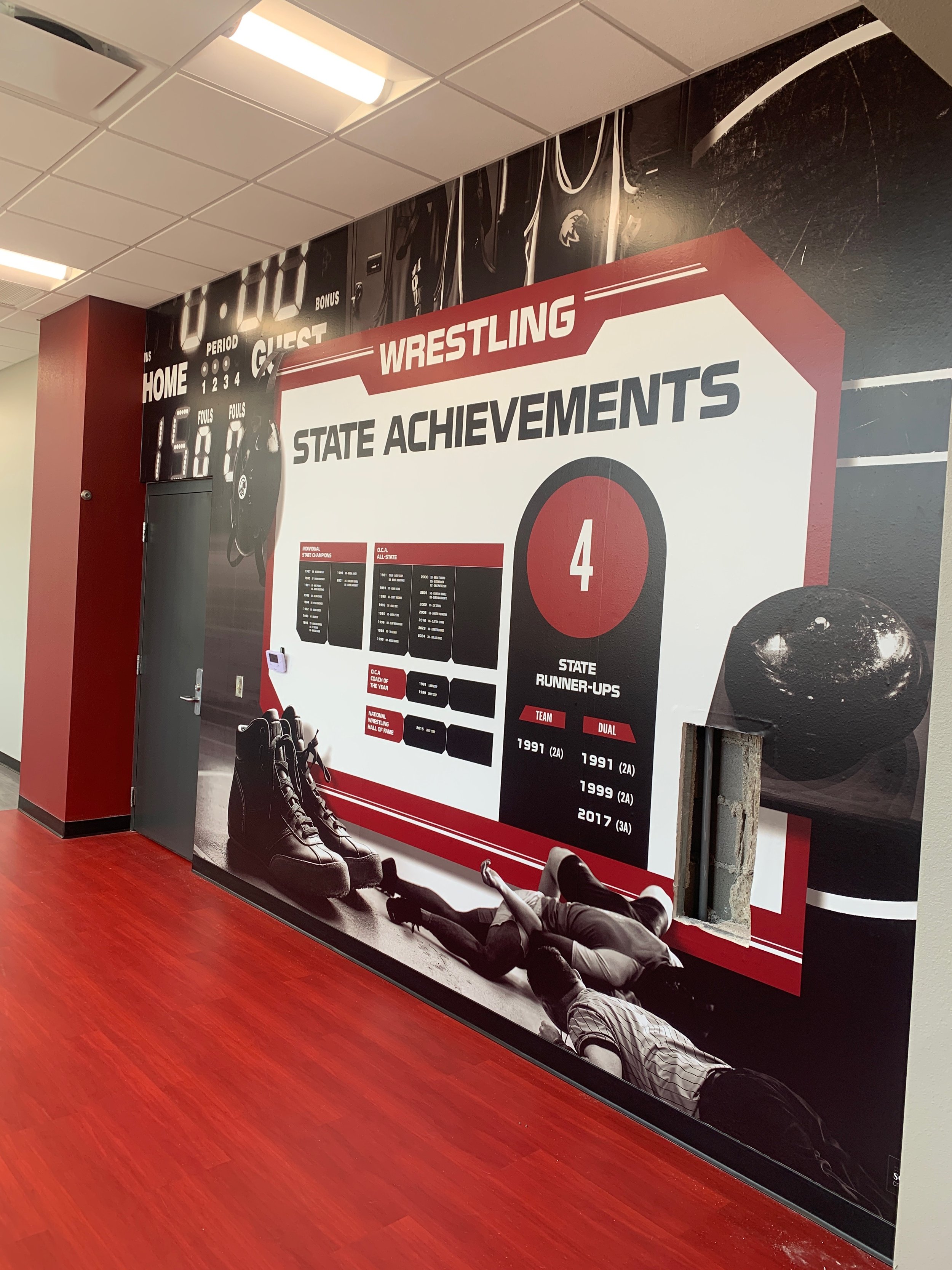 Wall mural honoring wrestling achievements at a school, featuring state runner-up titles, a pair of wrestling shoes, and a photograph of wrestlers on the mat.