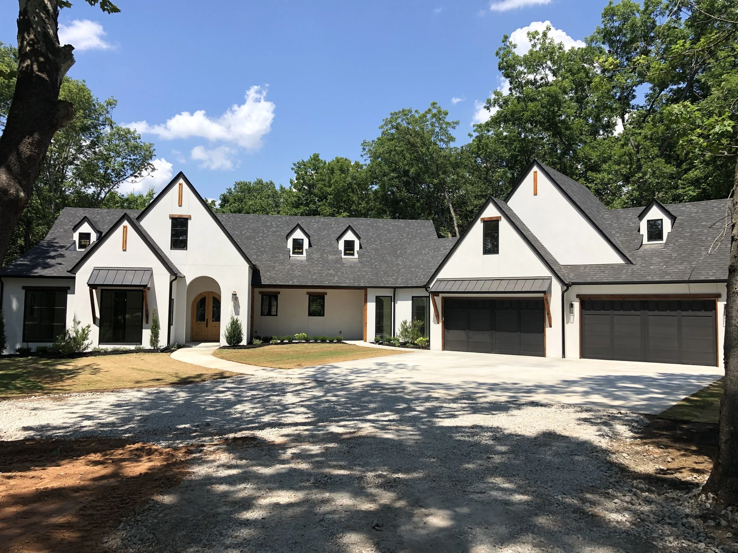 Modern white house with black roof, two-car garage, and landscaped front yard, surrounded by trees under a blue sky.