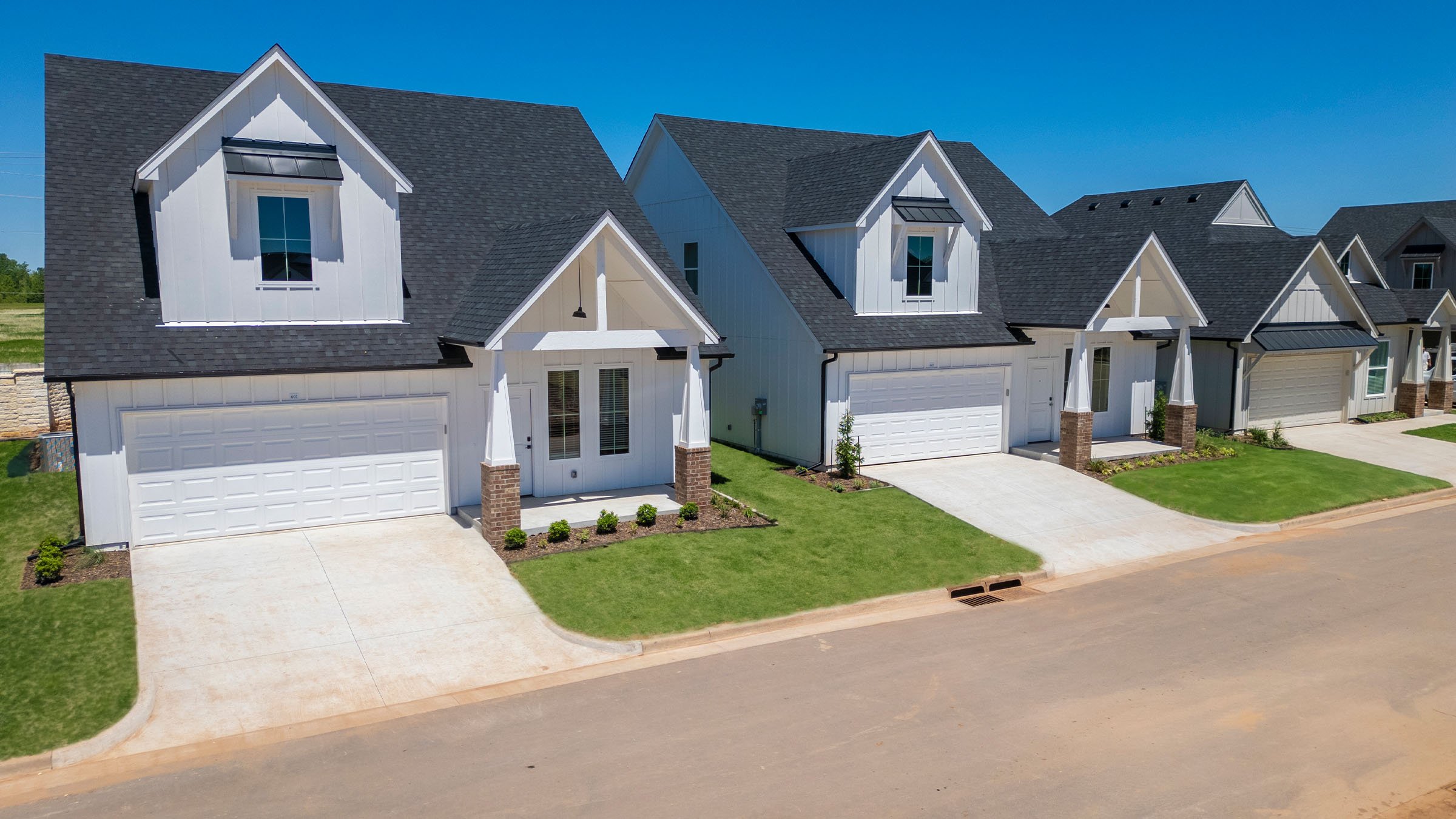 A row of modern two-story white houses with black roofs, attached garages, small front porches, and landscaped lawns under a bright blue sky.