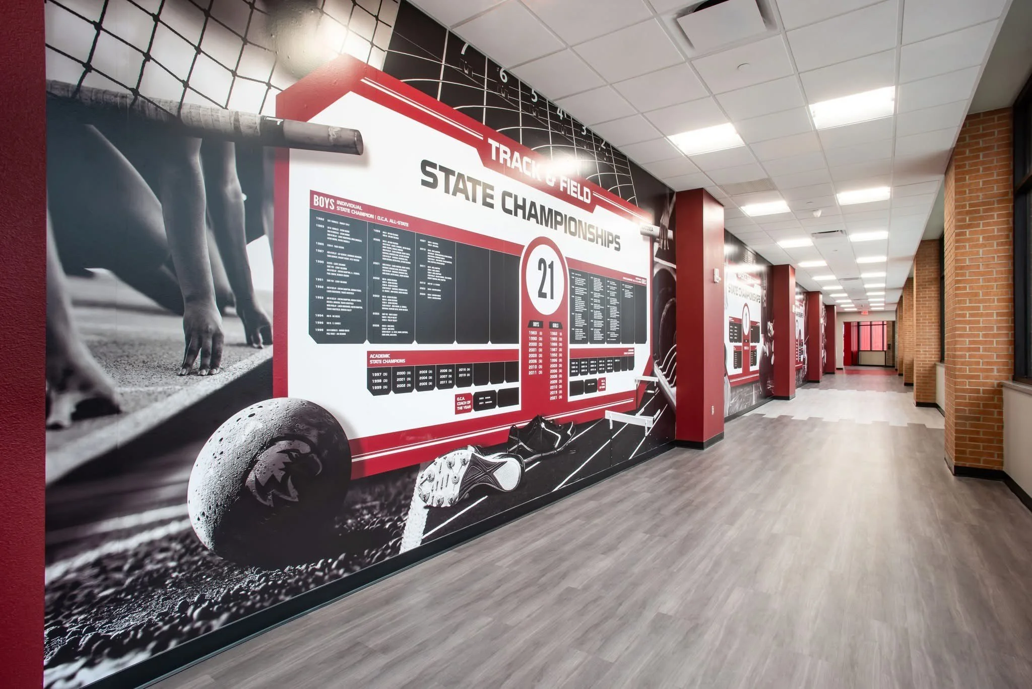 Indoor school hallway with wall graphics showing track and field state championships, with a large track graphic and text listing champions and years, and a corridor with brick walls and ceiling lights.