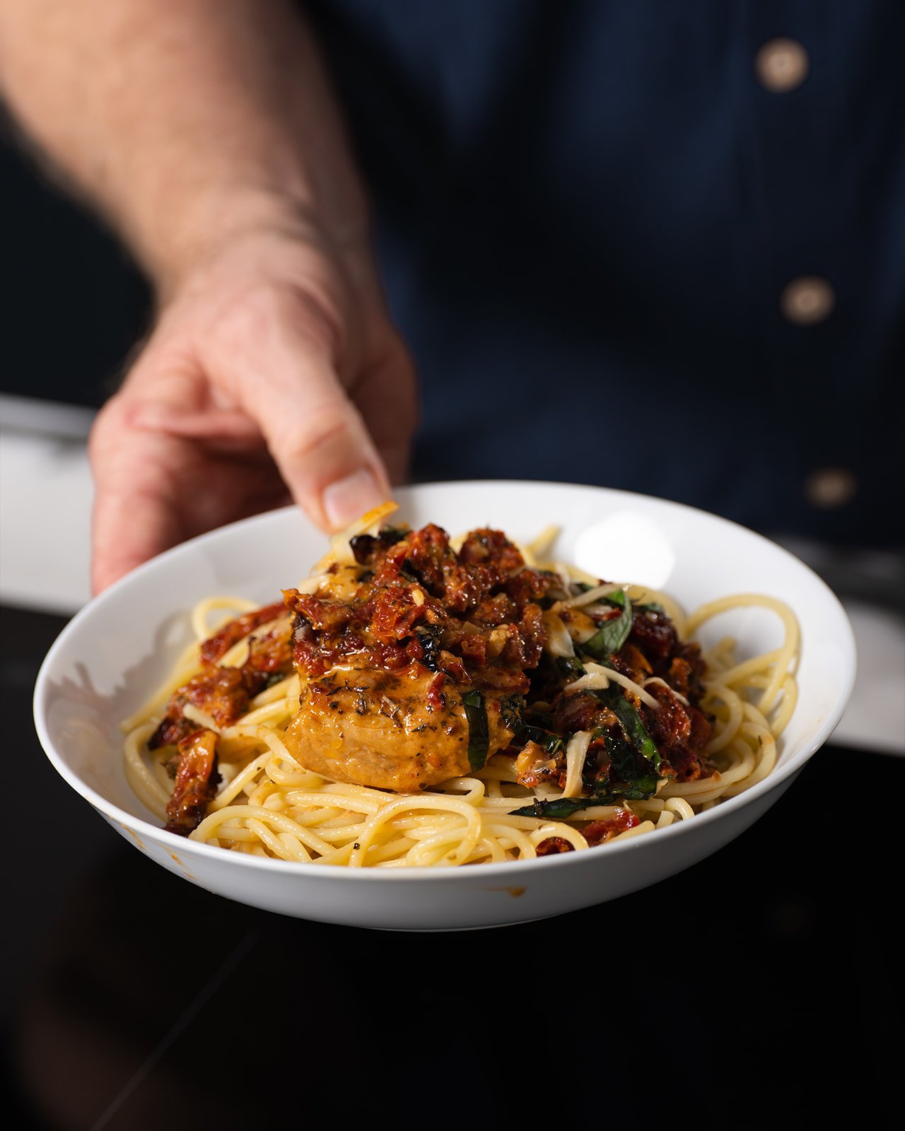a man's hand offering plated chicken over pasta in a white bowl