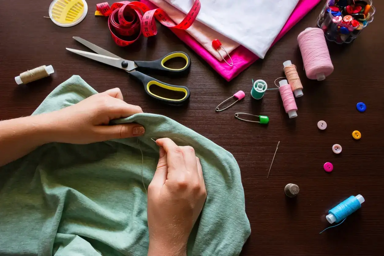 Person sewing fabric on a wooden table surrounded by sewing tools and supplies, including scissors, measuring tape, spools of thread, safety pins, buttons, and a container of sewing pins.