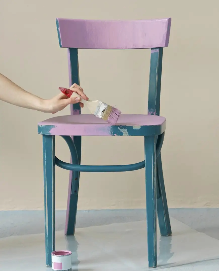 A person painting a wooden chair with pink and blue paint, using a brush.