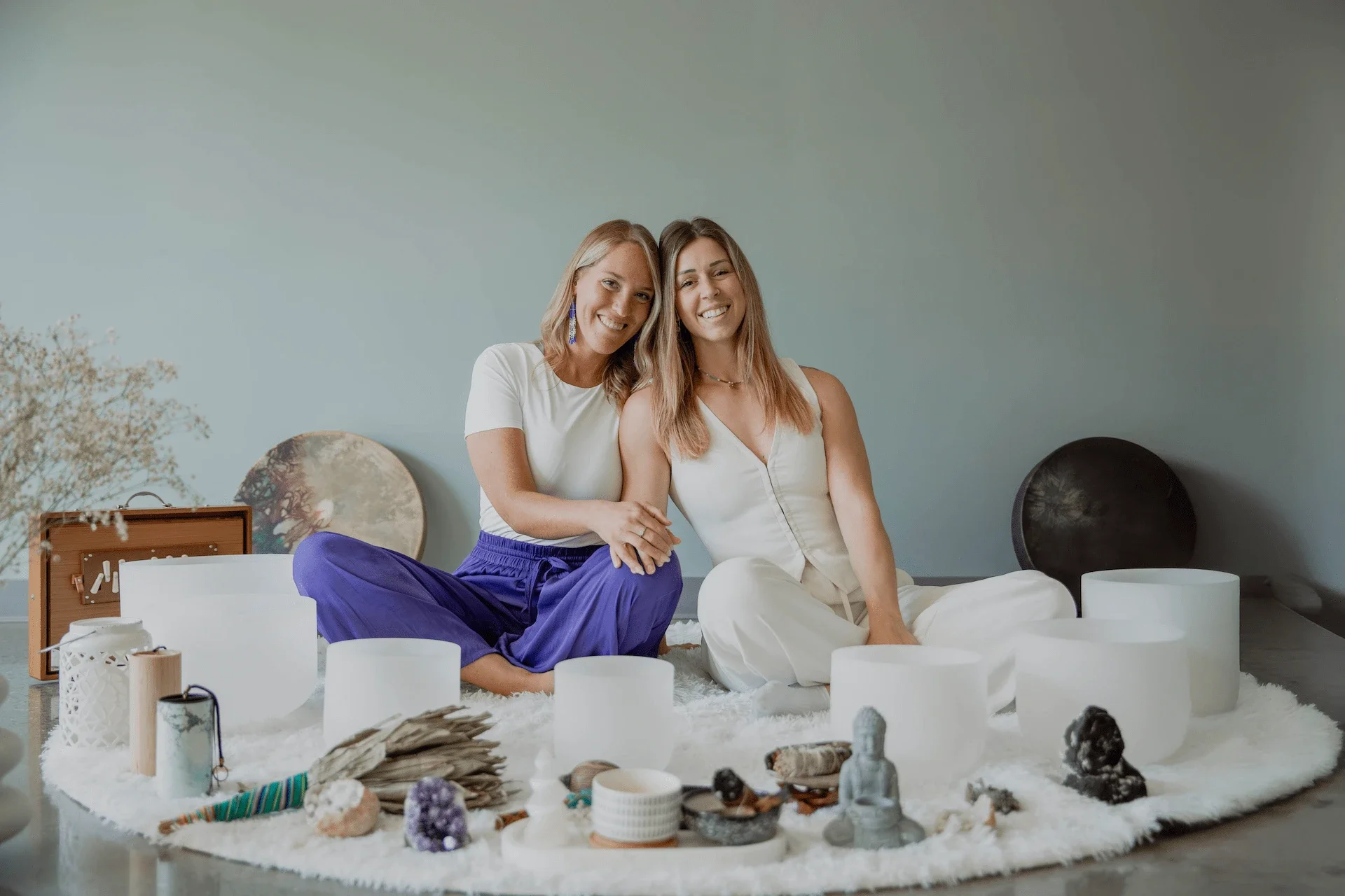 Deux femmes assises ensemble sur un tapis, entourées de bols chantants en cristal et d’objets spirituels, souriant à la caméra.