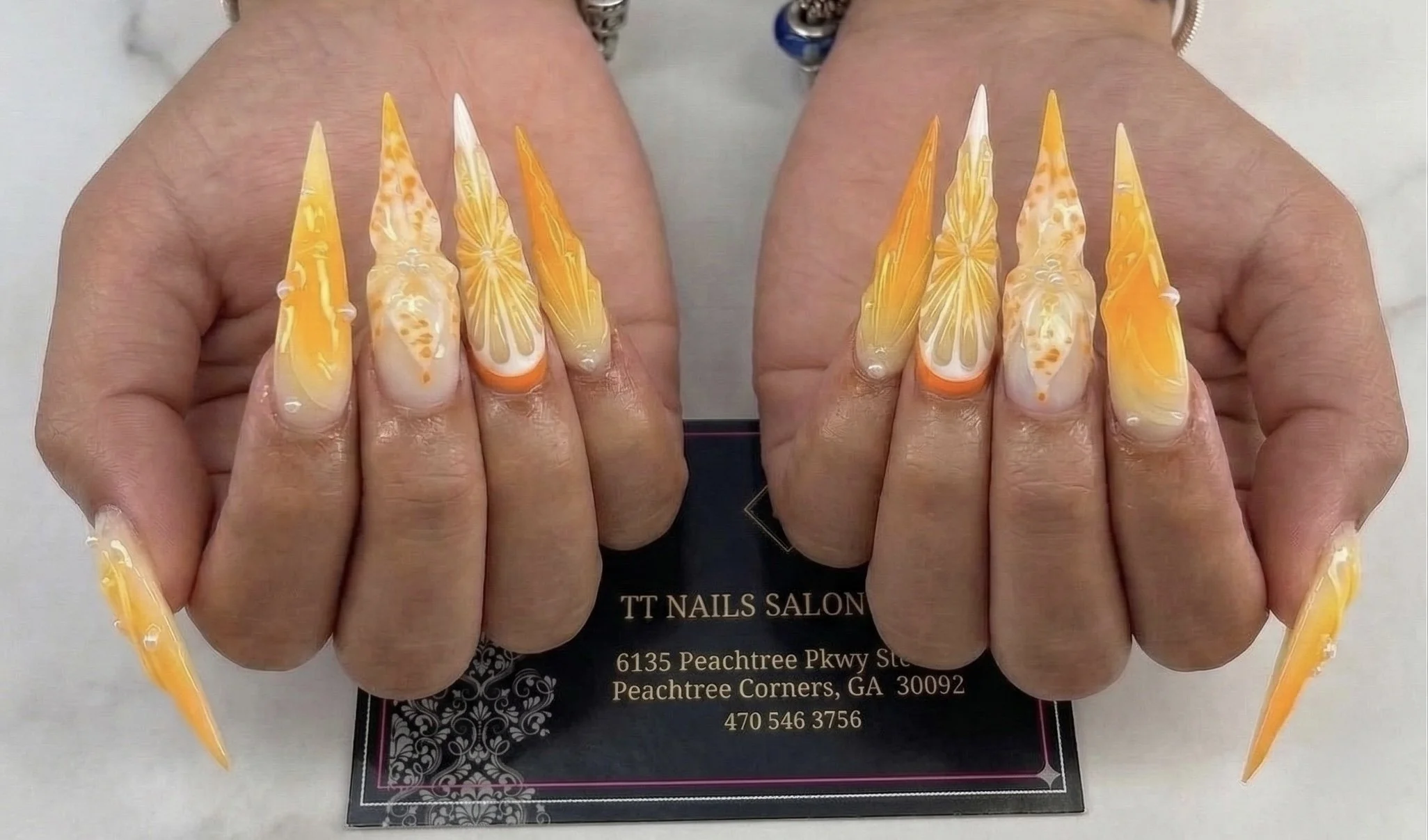 Long orange and yellow stiletto nail art with translucent floral designs, displayed on both hands.