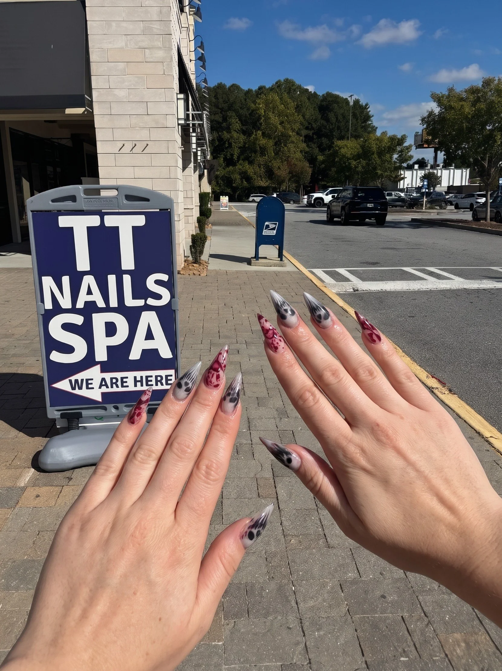 Hands with long decorated stiletto nails outside a nail spa, beside a sign reading “TT Nails Spa” and a USPS mailbox.