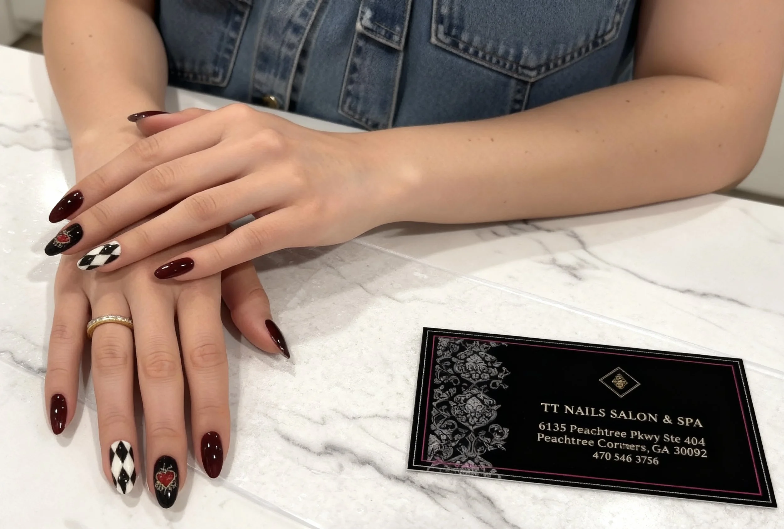 Person with manicured nails displaying a manicure with black and white checkered pattern and red heart accents, resting hands on a white marble countertop with a black business card from TT Nails Salon & Spa.