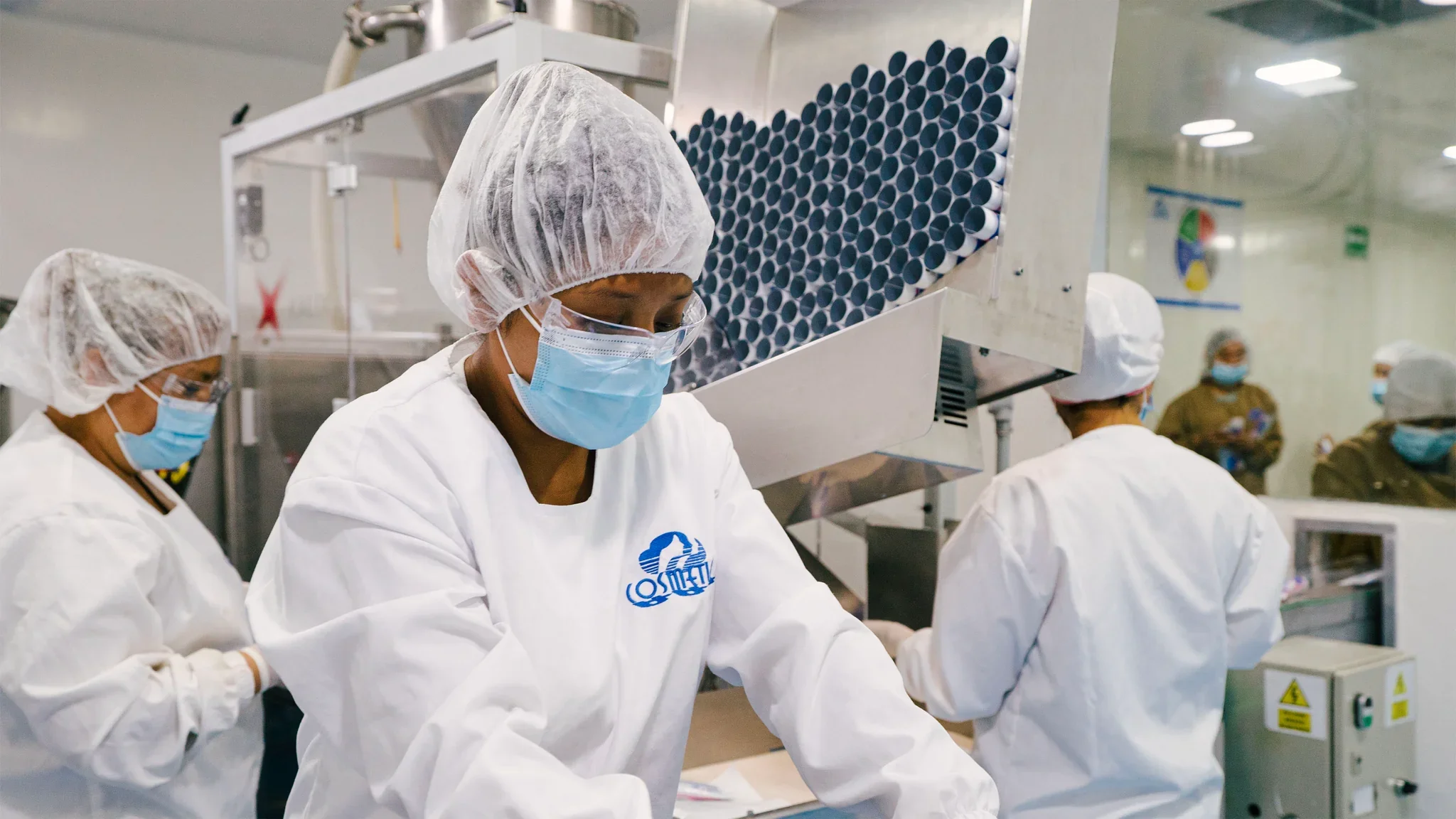 Workers wearing protective gear, including hair nets and face masks, working in a clean manufacturing environment with machinery and tubes.