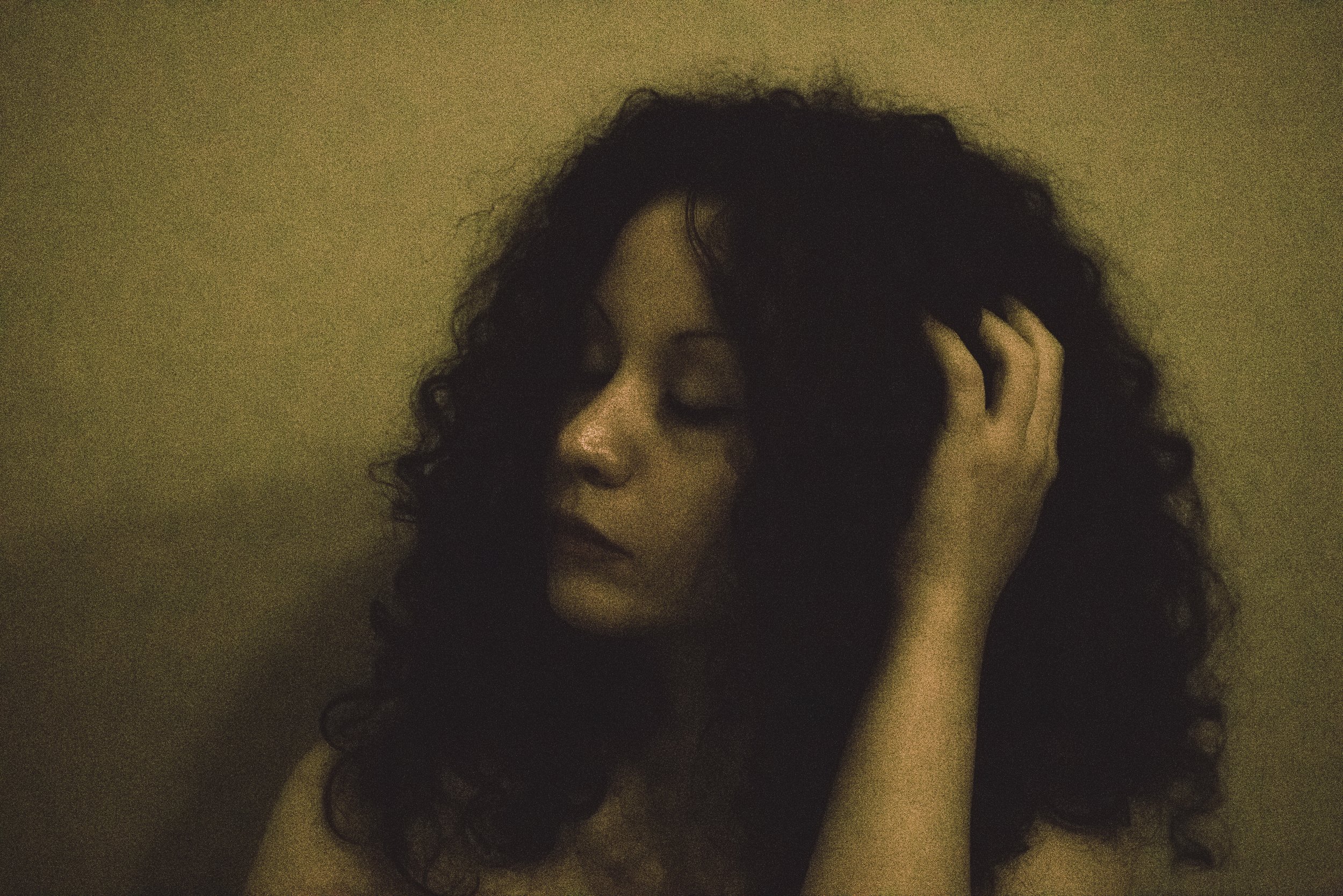 A woman with curly black hair touching her head with her eyes closed against a plain background.