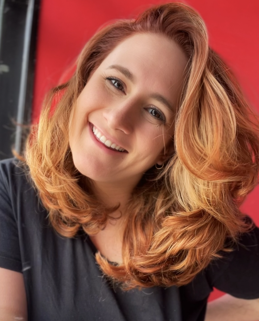 A smiling woman with wavy red hair posing in front of a red background.