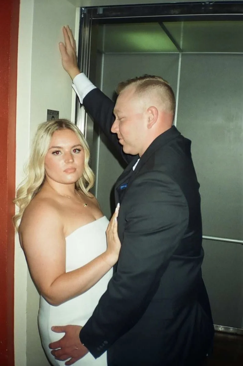 A couple standing in front of an elevator, with the woman in a strapless white dress and the man in a black suit, appearing close and intimate.