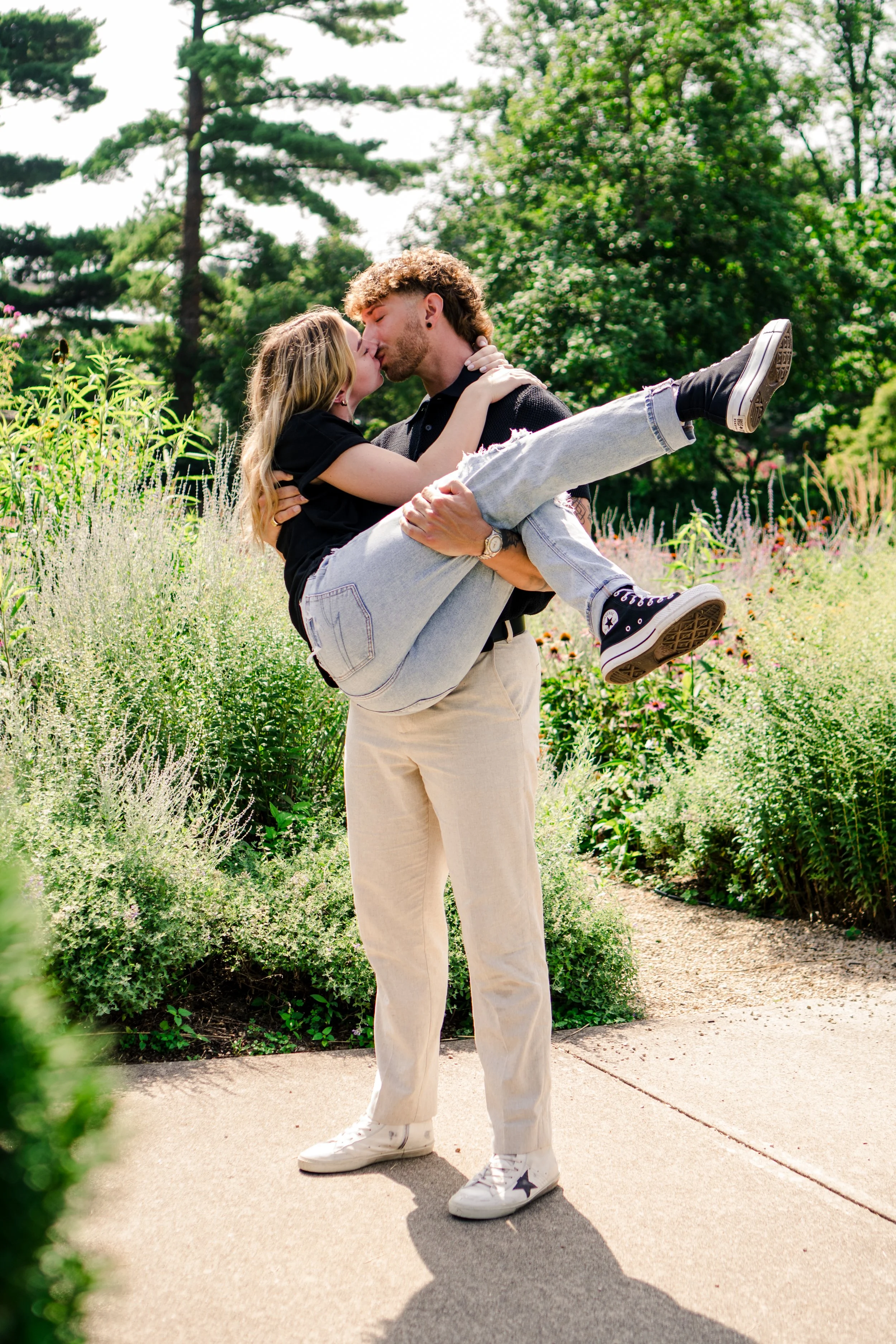 A man holding a young girl in his arms during a kiss in a lush, green park with tall trees and flowering plants.