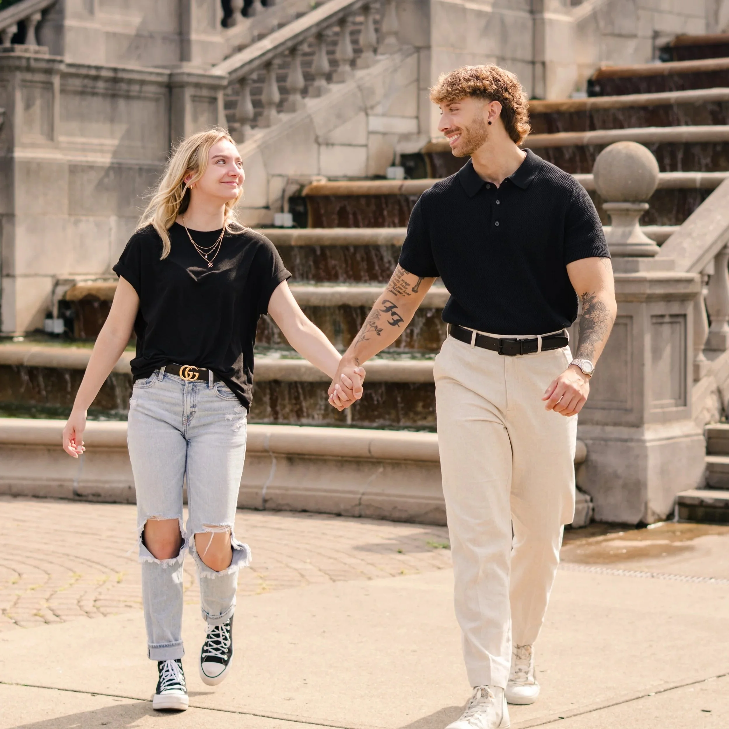 A young woman and a young man holding hands and walking outdoors near a stone fountain, smiling at each other.