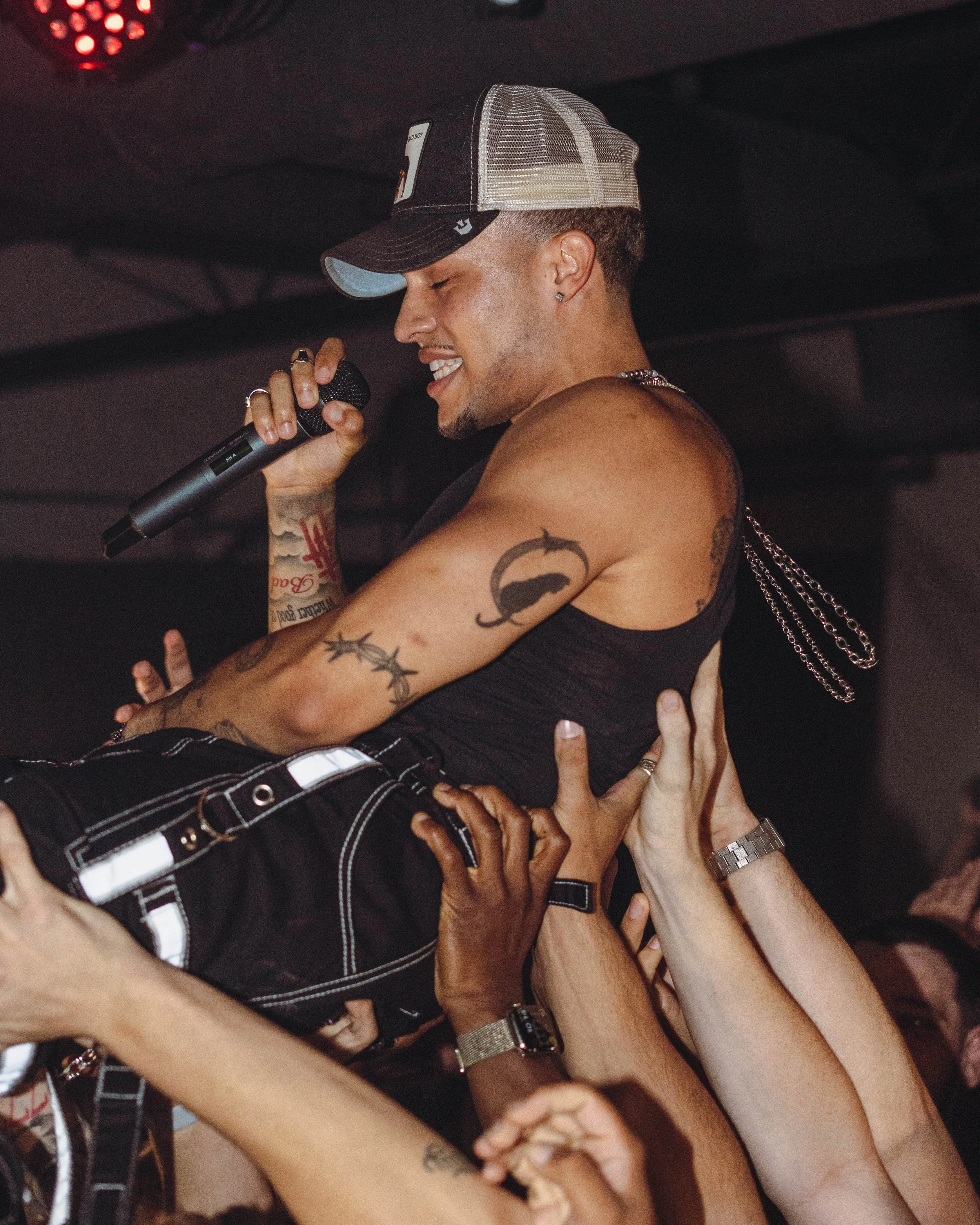A man with tattoos, wearing a black tank top and a trucker hat, is singing into a microphone while being lifted by a crowd at a concert or party event.