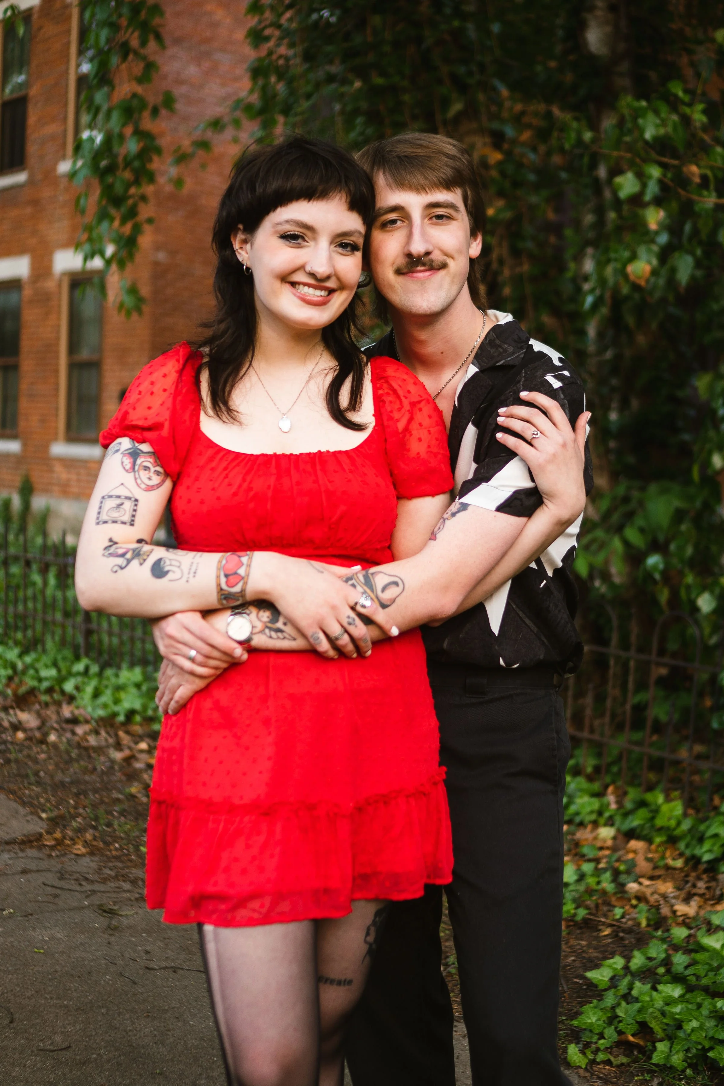 A happy couple embracing outdoors, woman in a red dress with tattoos on her arms and a tattoo on her thigh, man in a black and white patterned shirt, background of green plants and a brick building.