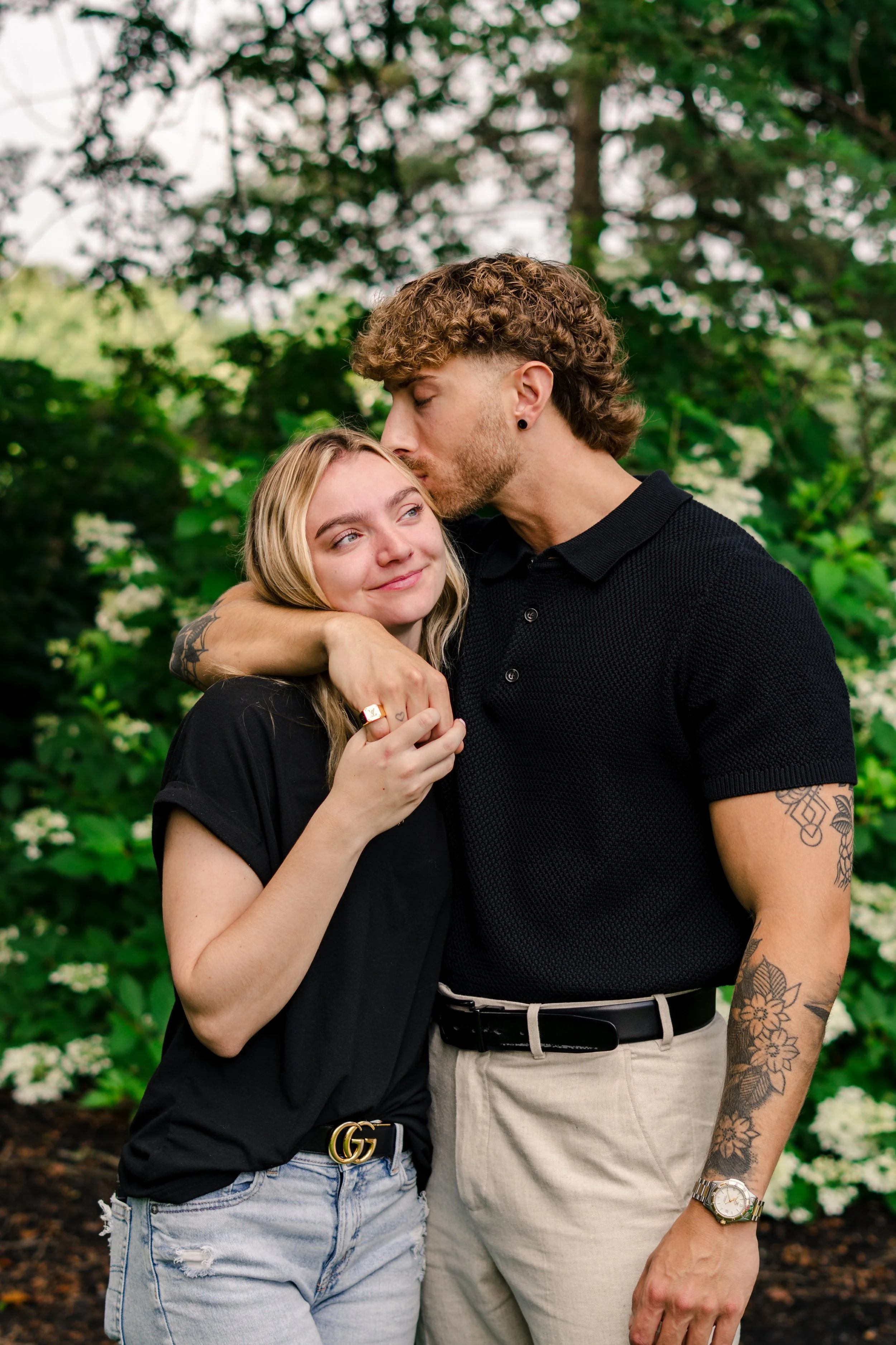 A young man is kissing a young woman on the forehead while hugging her. They are outdoors surrounded by green trees and bushes.