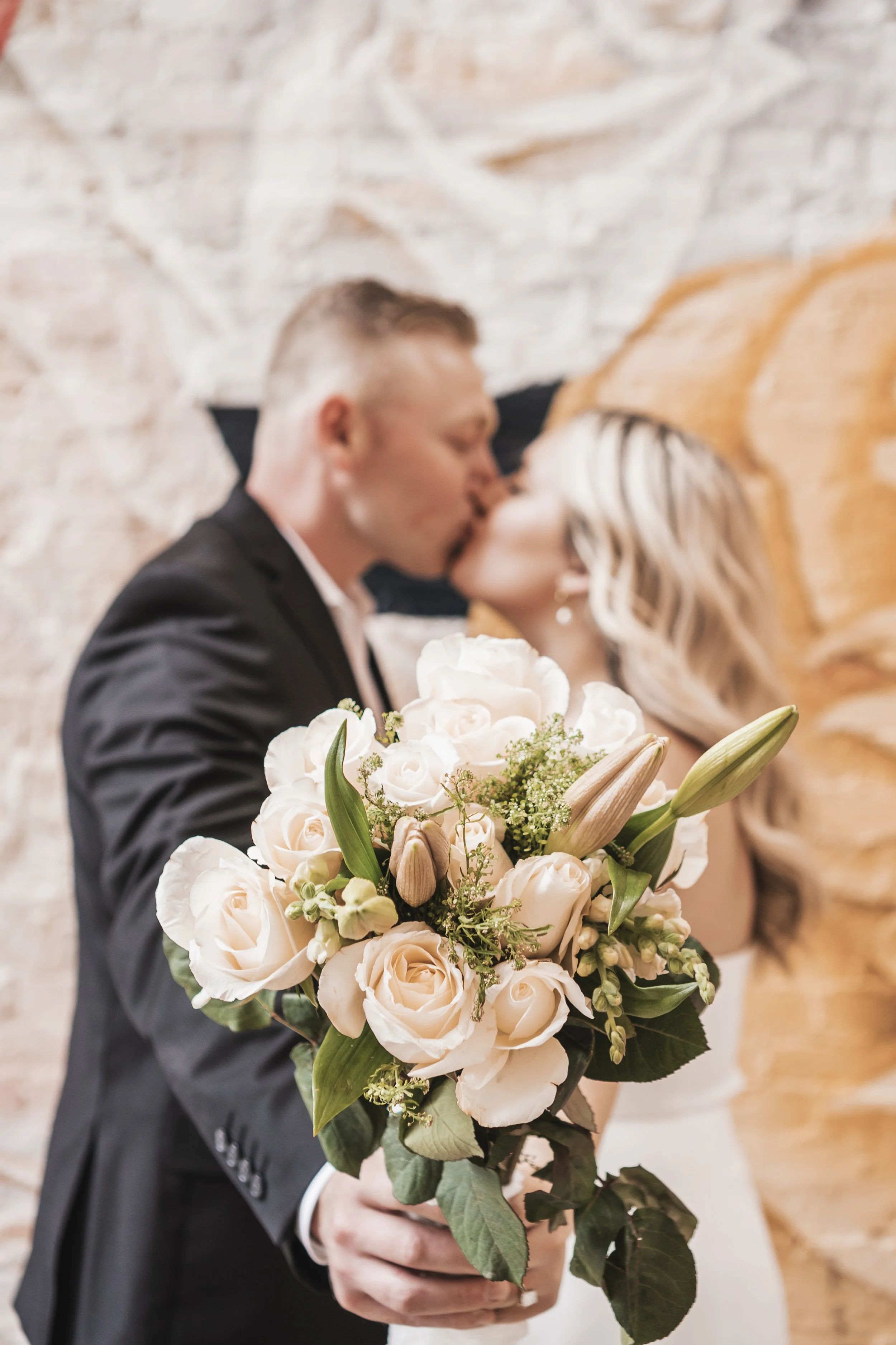 A bride and groom kissing behind a bouquet of pale pink roses and white lilies.