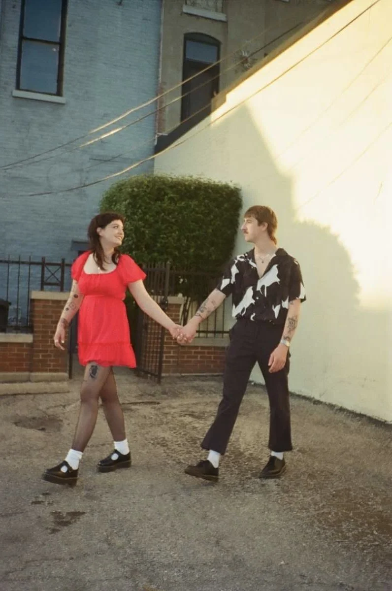 Two young women holding hands and walking outdoors at sunset or in a shaded area, with one wearing a red dress and the other wearing a black and white patterned shirt and black pants.