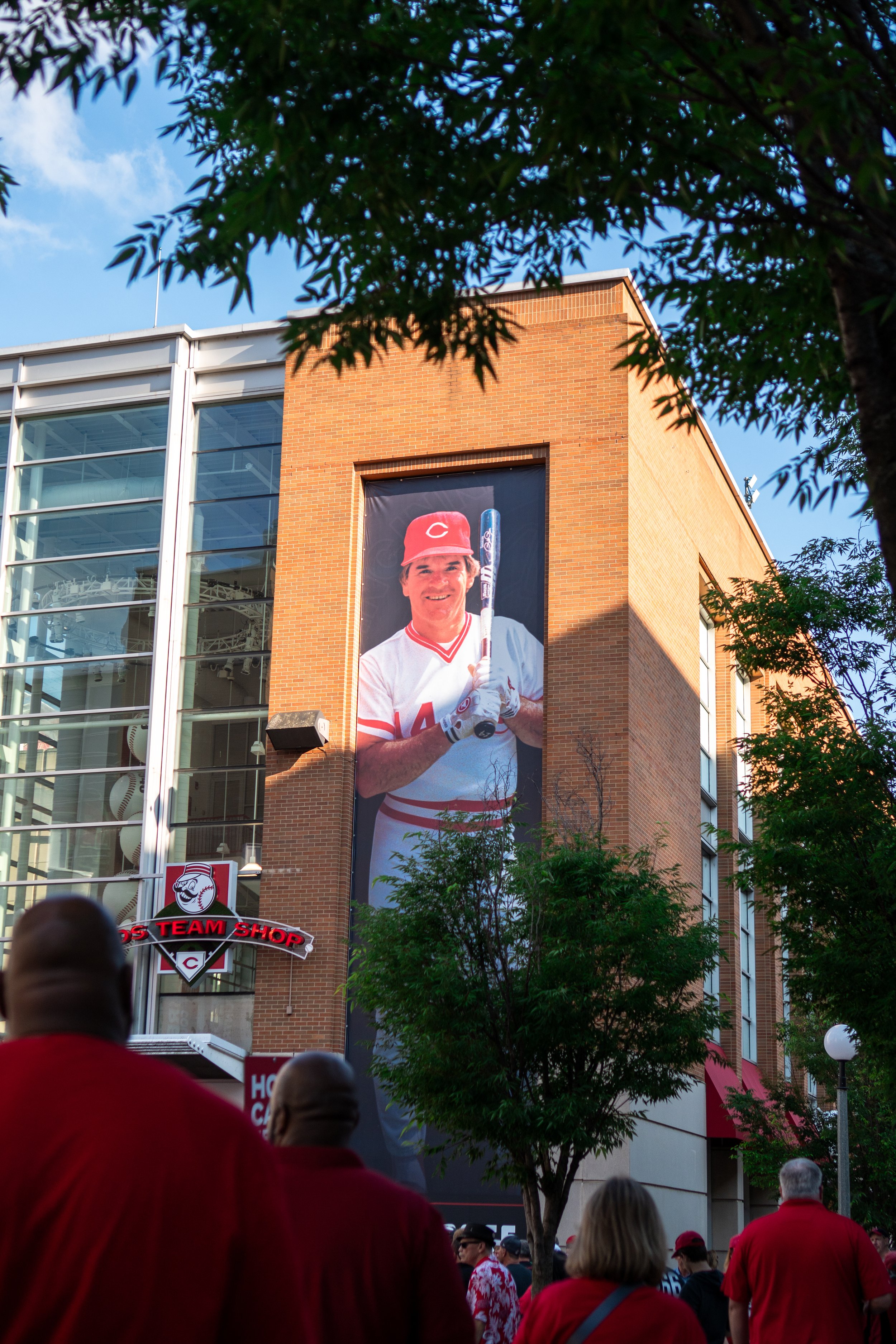 Crowd of people in red clothing looking at a large banner on a brick building, featuring a smiling baseball player in uniform holding a bat.