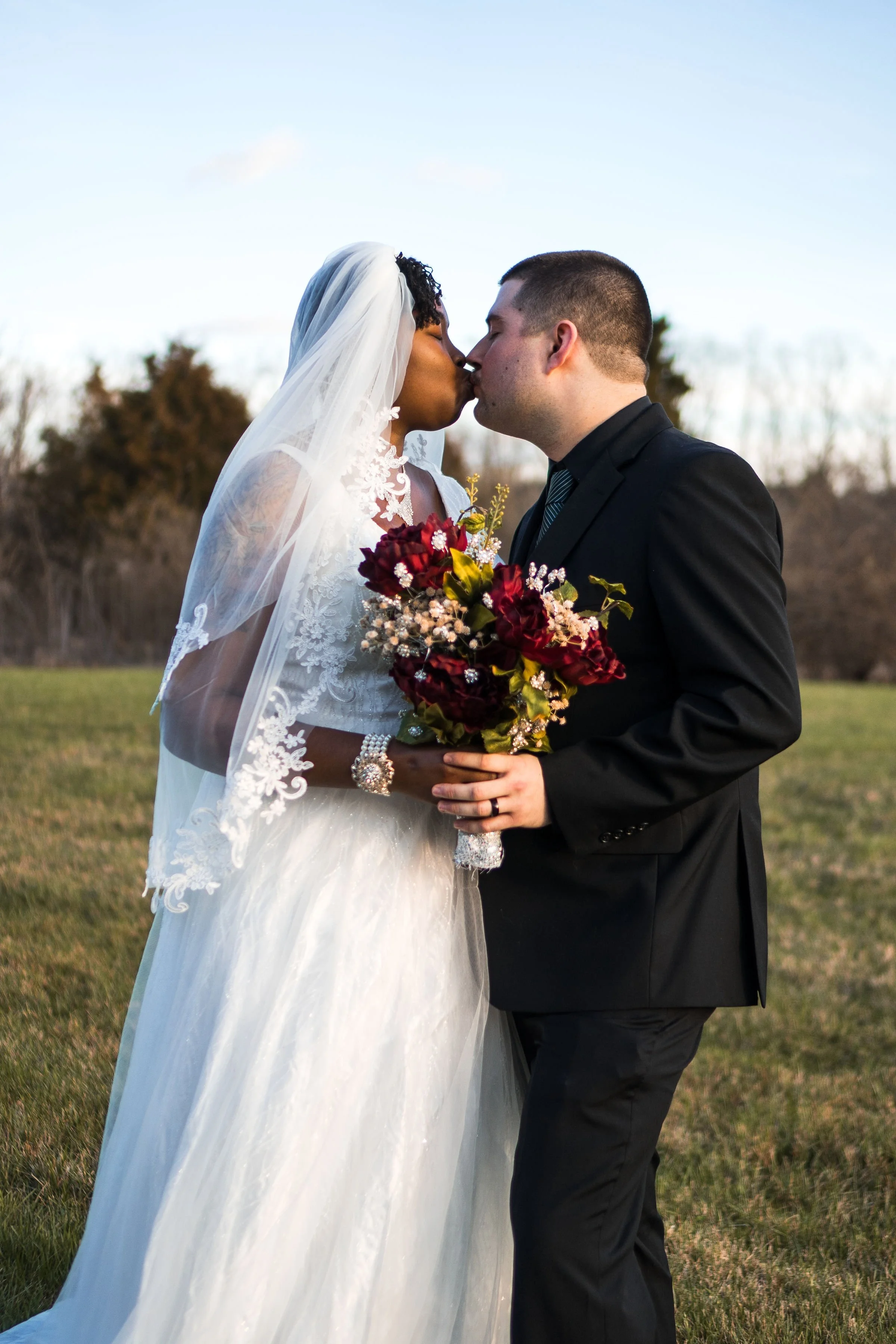 A bride and groom share a kiss outdoors on their wedding day, with the bride holding a bouquet of red flowers and wearing a lace wedding gown and veil, while the groom is dressed in a black suit and tie.