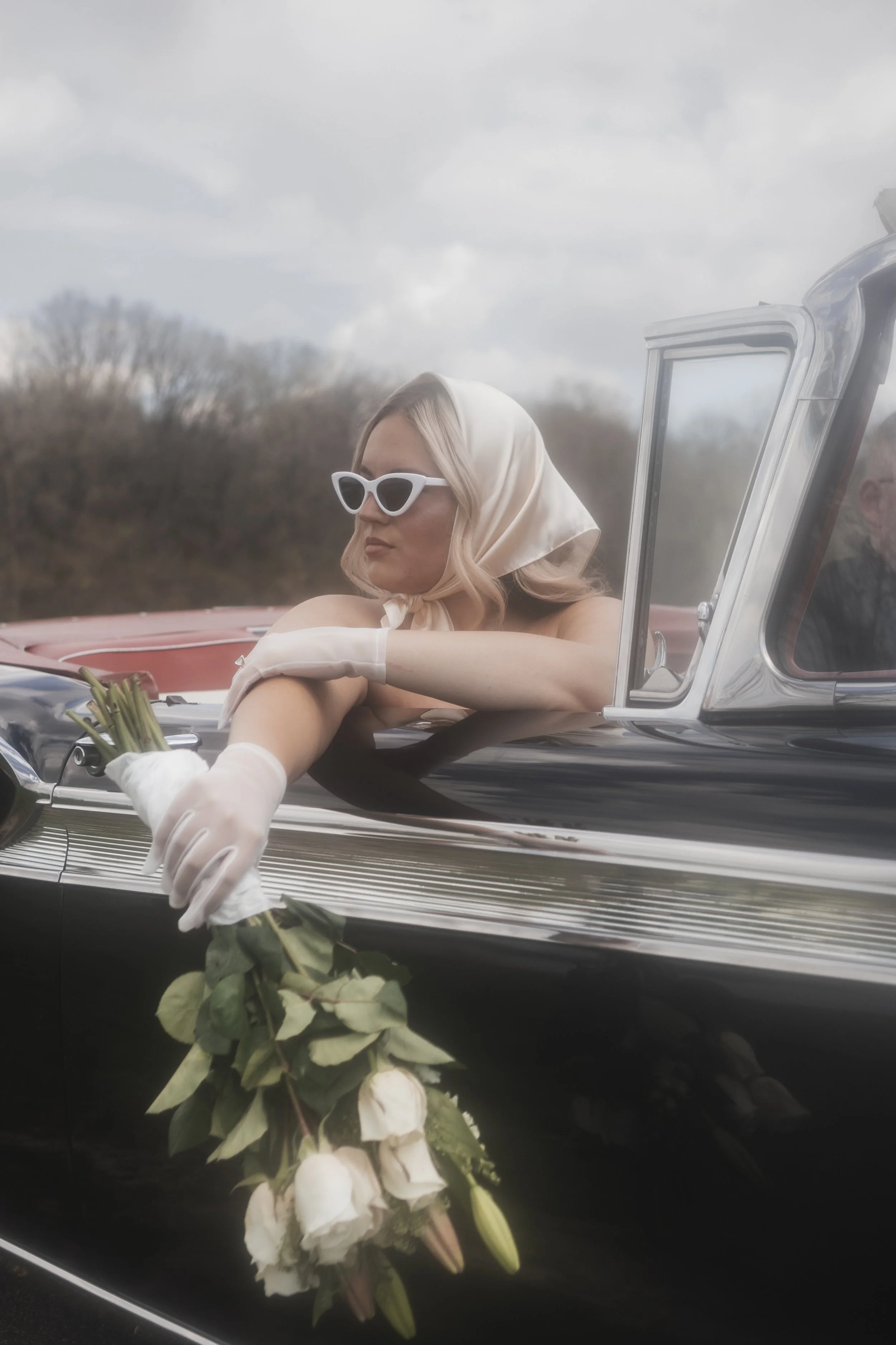 A woman with blonde hair, wearing white sunglasses, a headscarf, and white gloves, is sitting in a vintage black car with a bouquet of white roses hanging from the side, against a cloudy outdoor background.