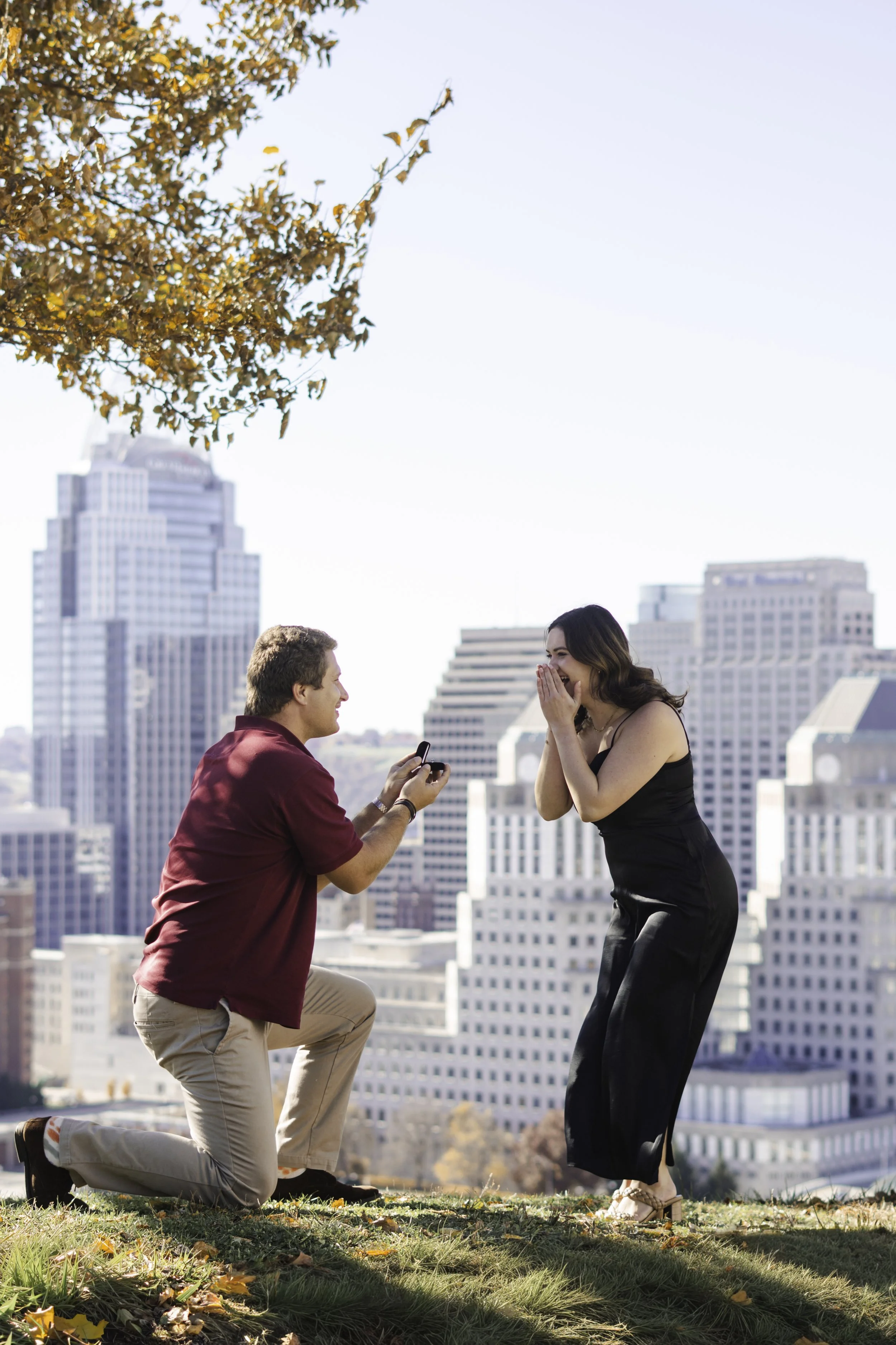 A man on one knee proposing to a woman on a grassy area with city buildings in the background. The woman is covering her mouth in surprise, and the man is holding a small box with a ring inside.