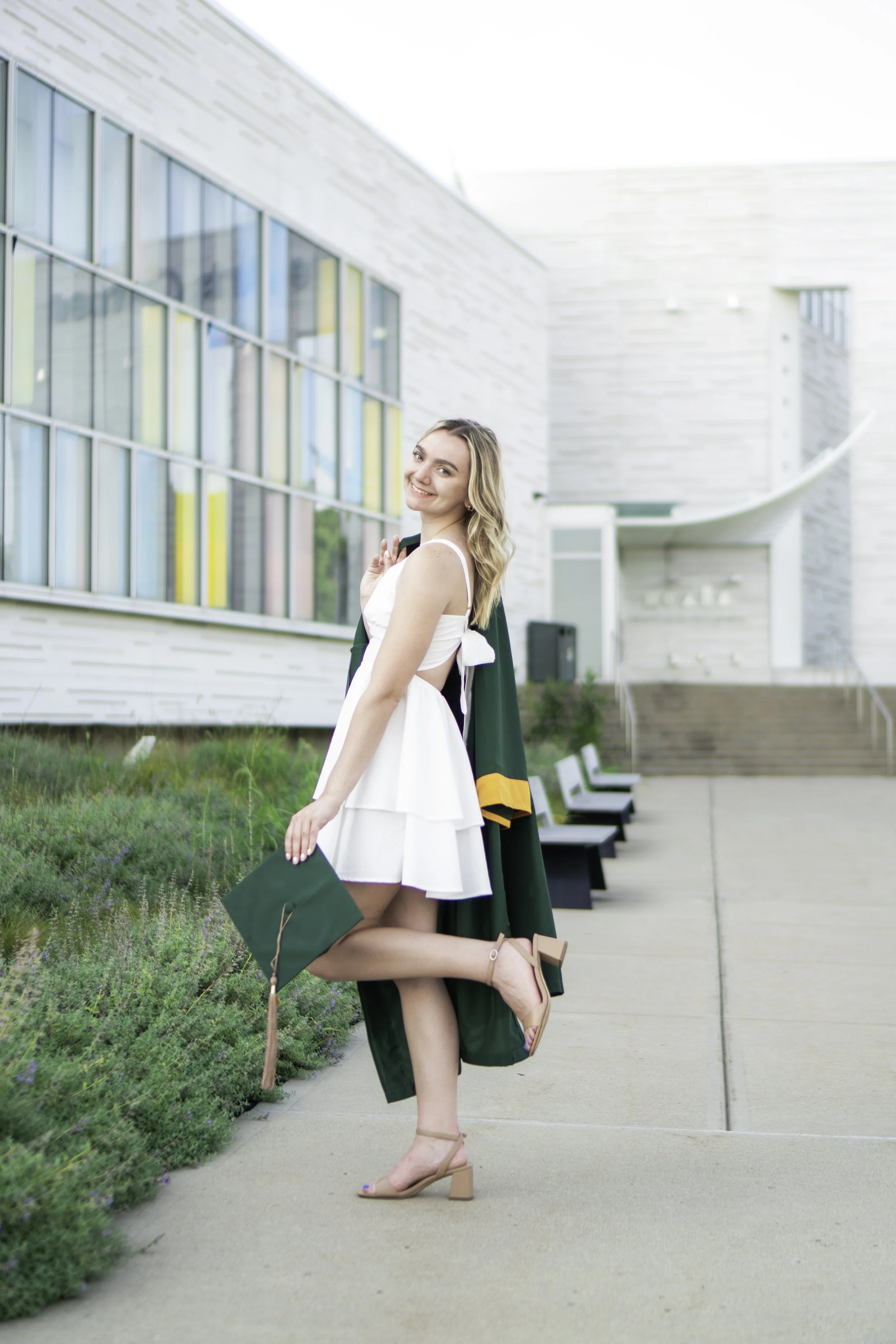 Young woman in white dress and heels celebrating graduation outdoors holding a diploma and wearing a graduation gown behind her