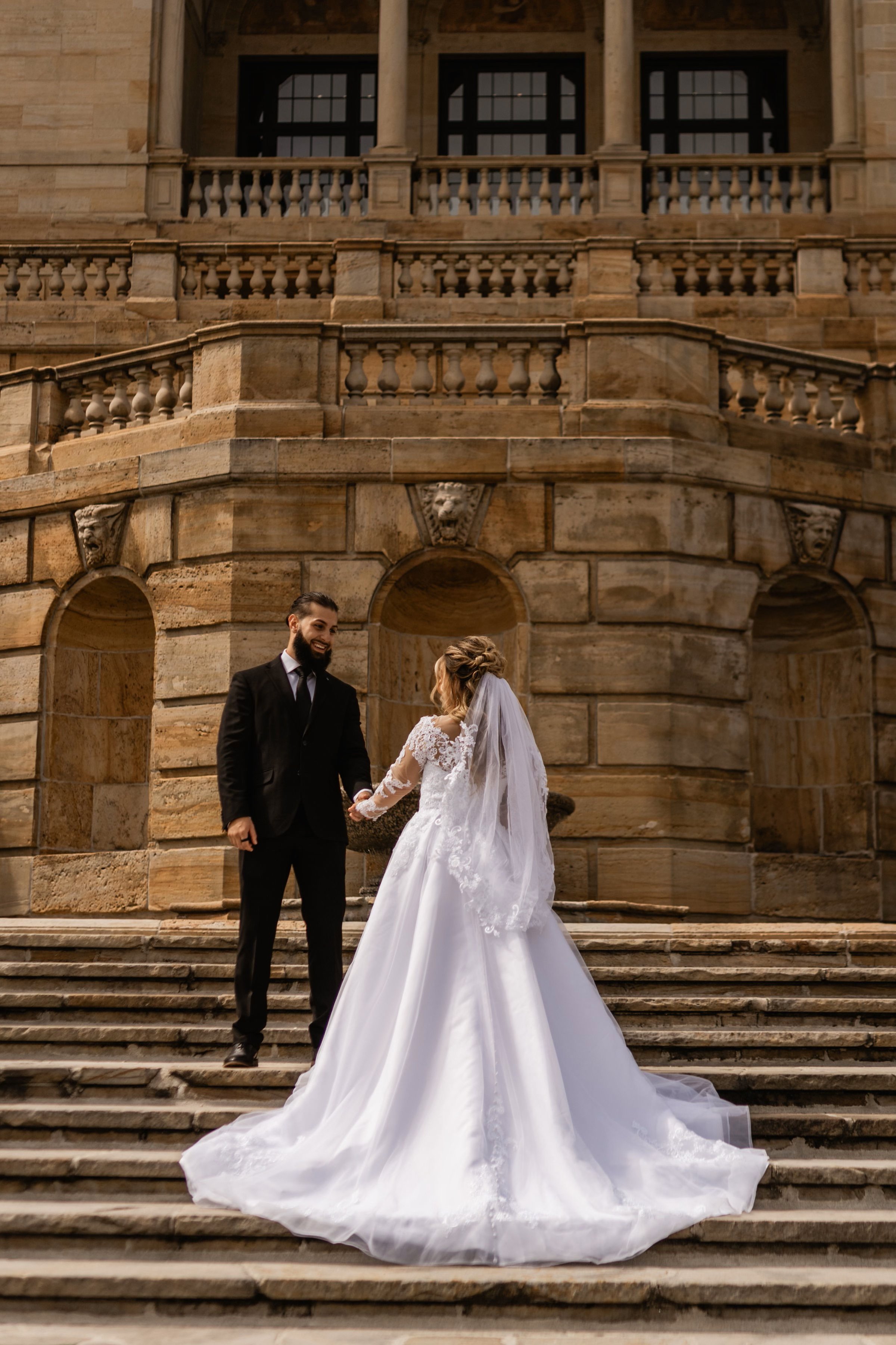 A bride and groom standing on stone steps outside a historic building, holding hands and smiling at each other during their wedding.