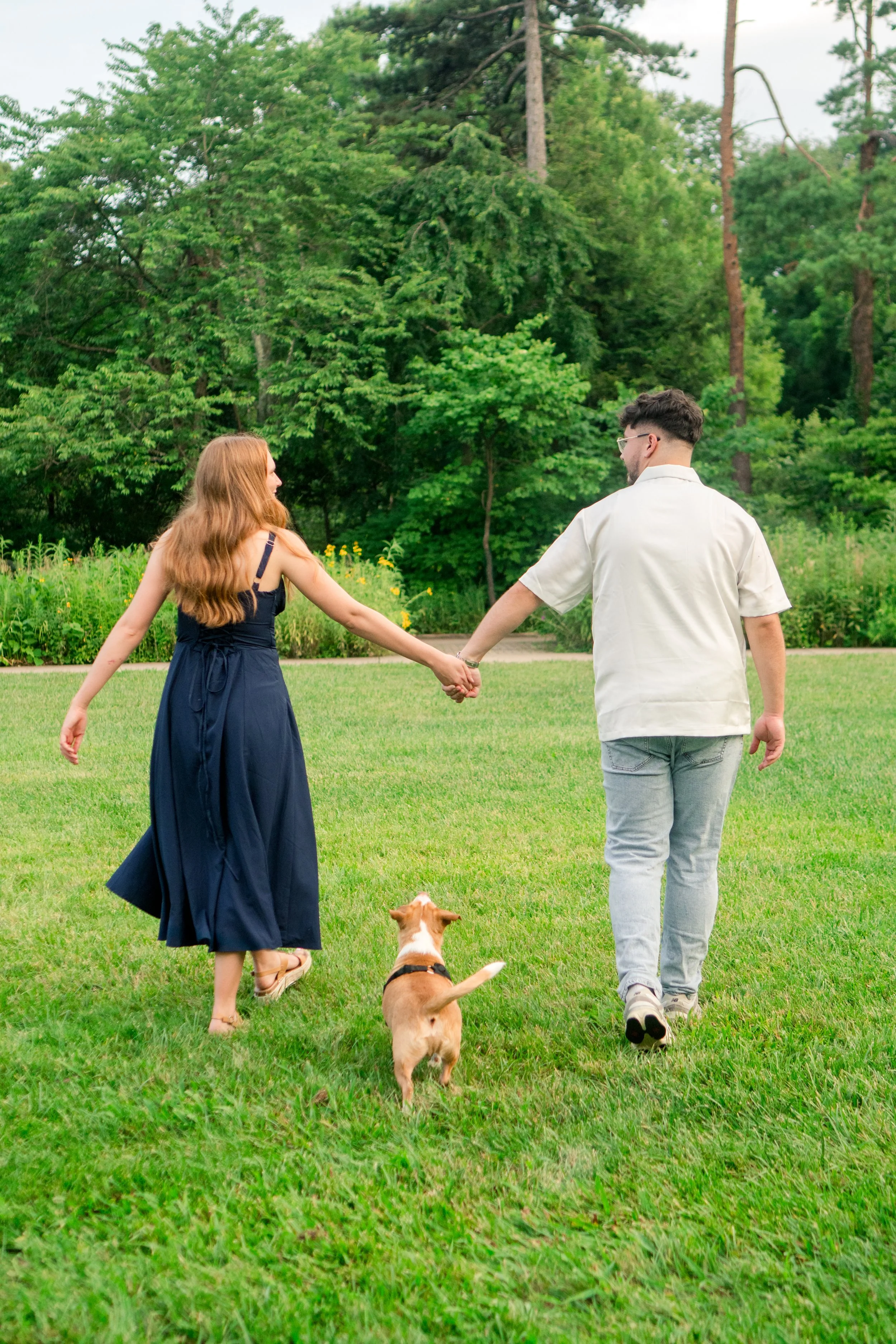 A young couple walking hand in hand in a park with a small dog, green grass, and trees in the background.