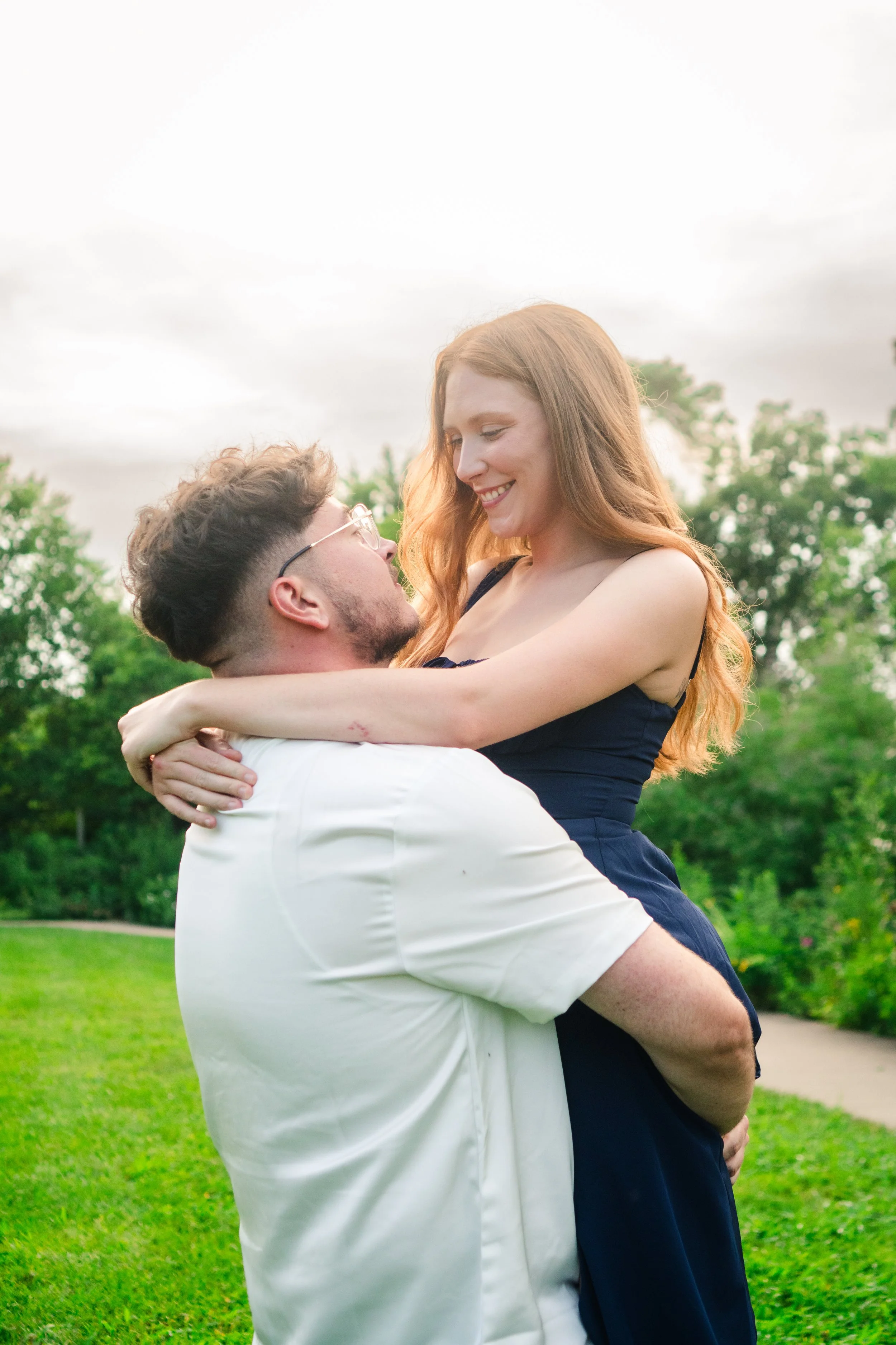 A happy couple outdoors, with the man holding the woman in his arms. The woman is smiling down at the man, and they are surrounded by green trees and grass, with an overcast sky overhead.