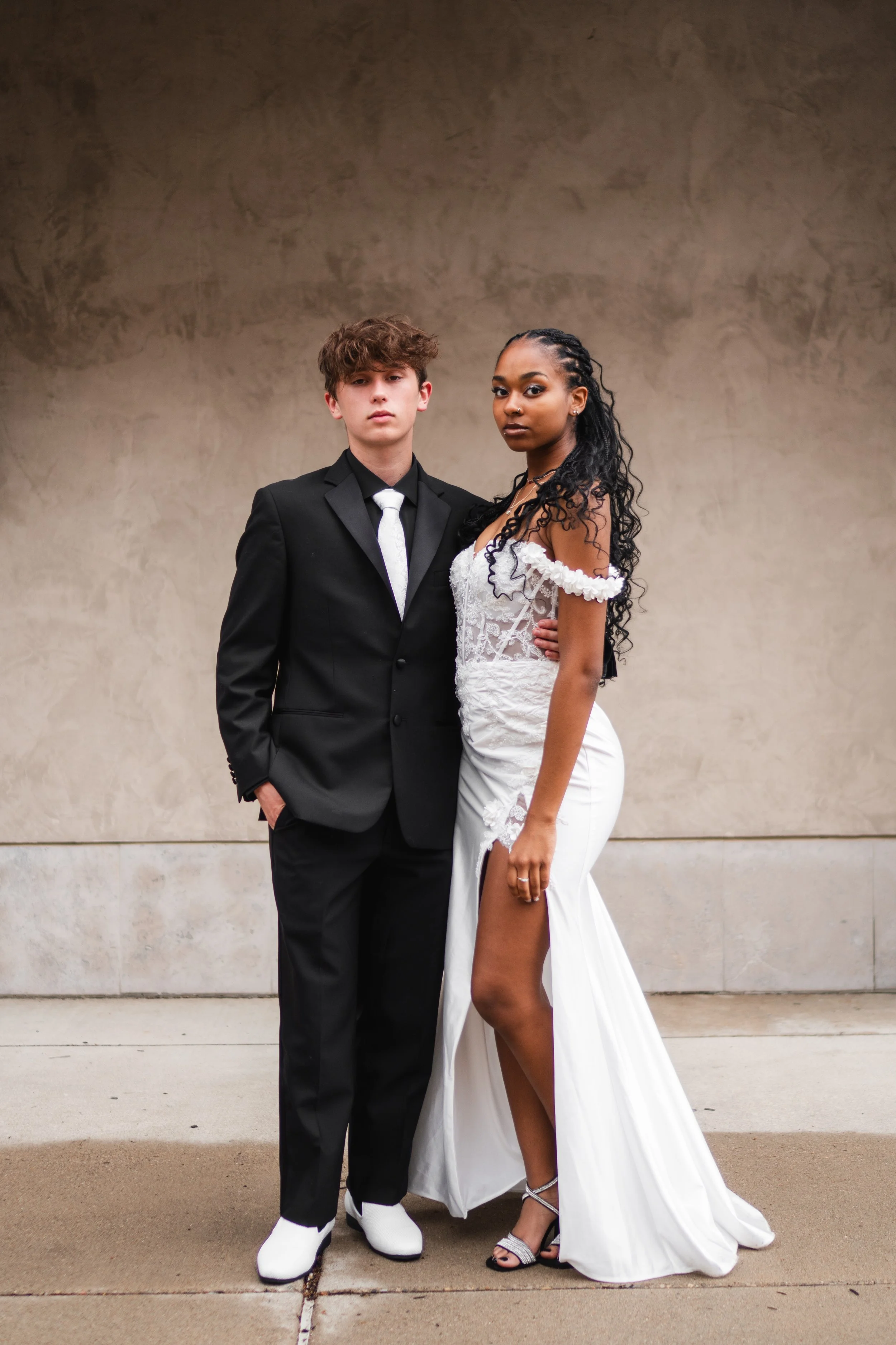 A young couple in formal attire standing close together, the man in a black suit with a white tie and the woman in a white dress with lace details, on a city sidewalk against a plain wall background.