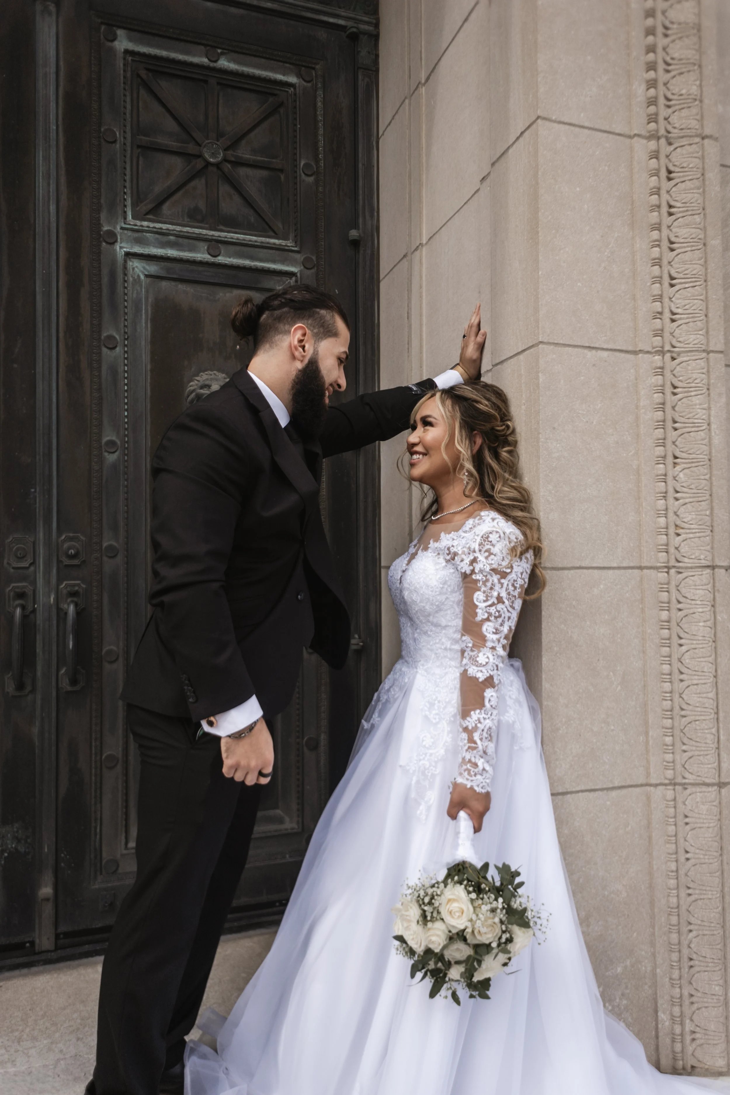 A bride and groom on their wedding day, standing close to a stone wall and black door. The groom, dressed in a black suit, is leaning with his hand against the wall, smiling at the bride. The bride, in a white lace wedding gown, is holding a bouquet of white roses and smiling back. They are in an affectionate pose, capturing a romantic moment.