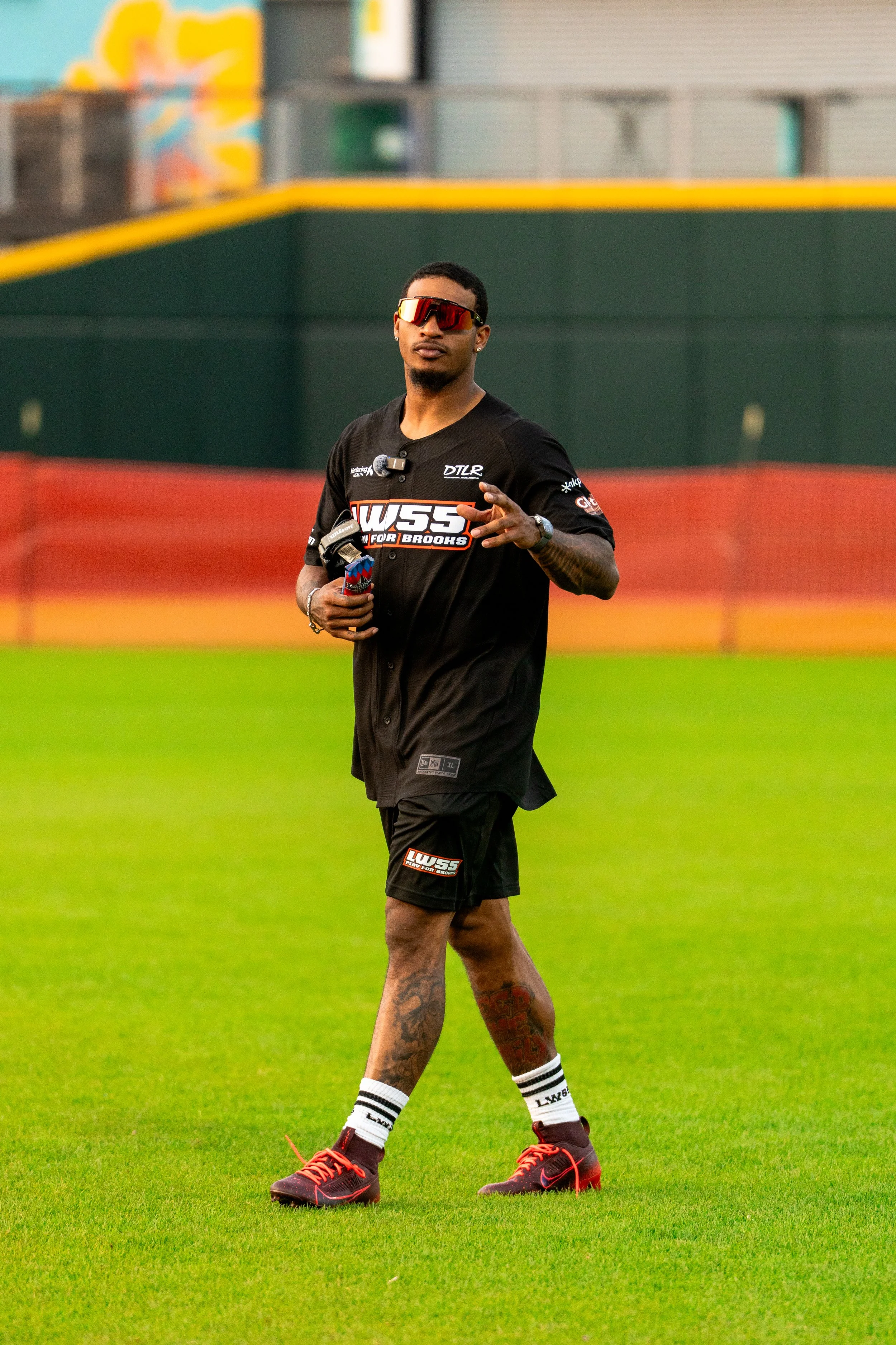 A male athlete on a baseball field wearing black sports attire with sponsor logos, red and black cleats, white socks with black stripes, and red sports sunglasses, holding a water bottle, standing on bright green grass.