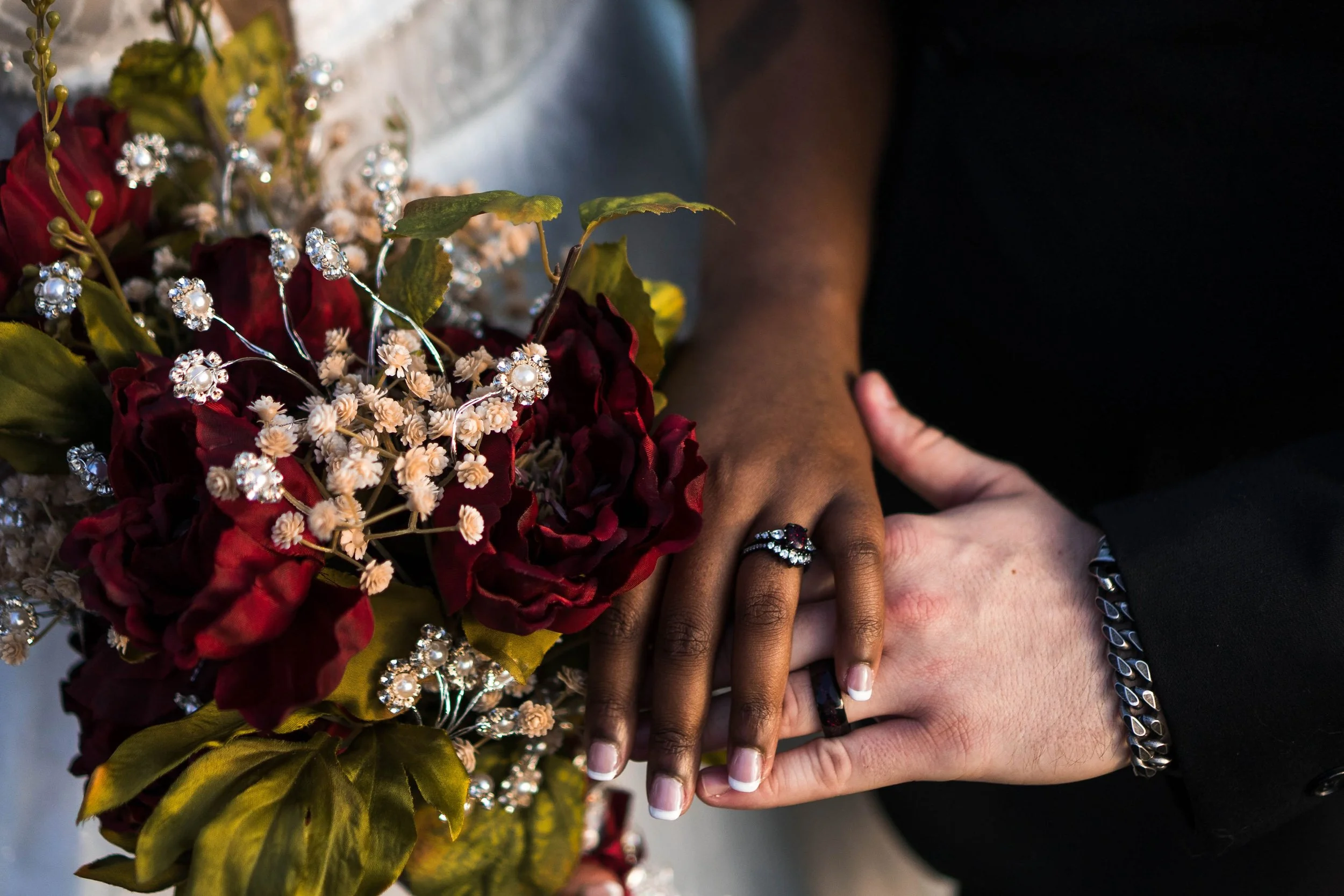 Close-up of newlywed couple's hands with wedding rings, holding a bouquet of dark red roses, baby's breath, pearl and rhinestone jewelry, and greenery.