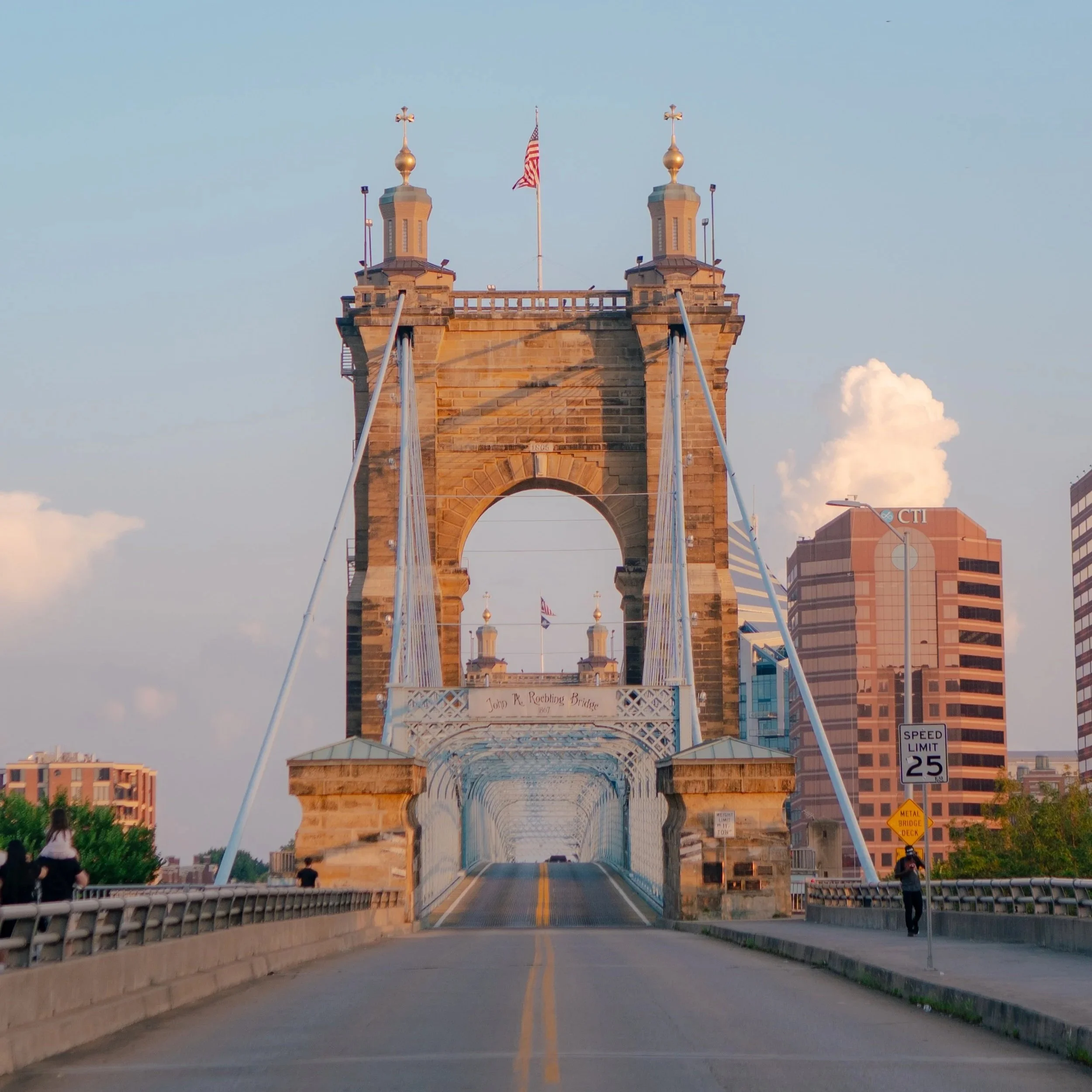 The John R. Rebling Bridge, a historic suspension bridge in Cincinnati, Ohio, with a view of downtown buildings in the background, a few pedestrians, and a clear sky.