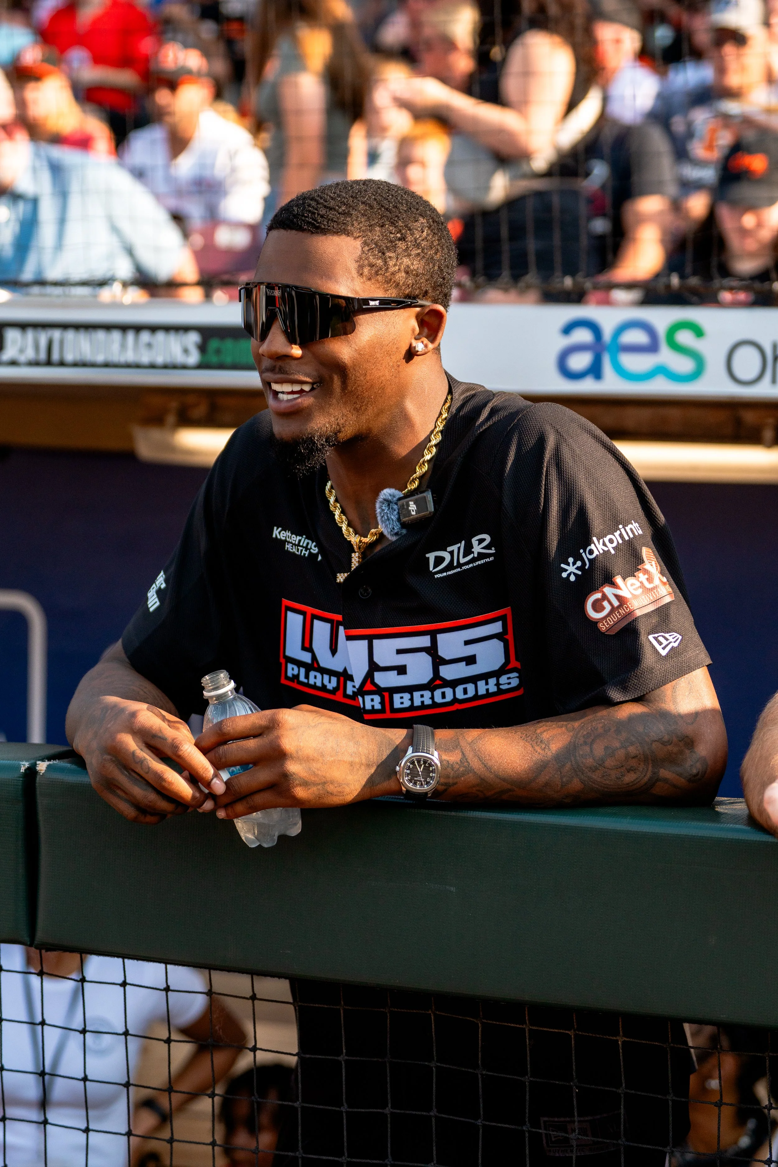 A man wearing sunglasses, a black T-shirt with a logo, gold chain, and watch, standing behind a dugout at a sports event, holding a water bottle and smiling.