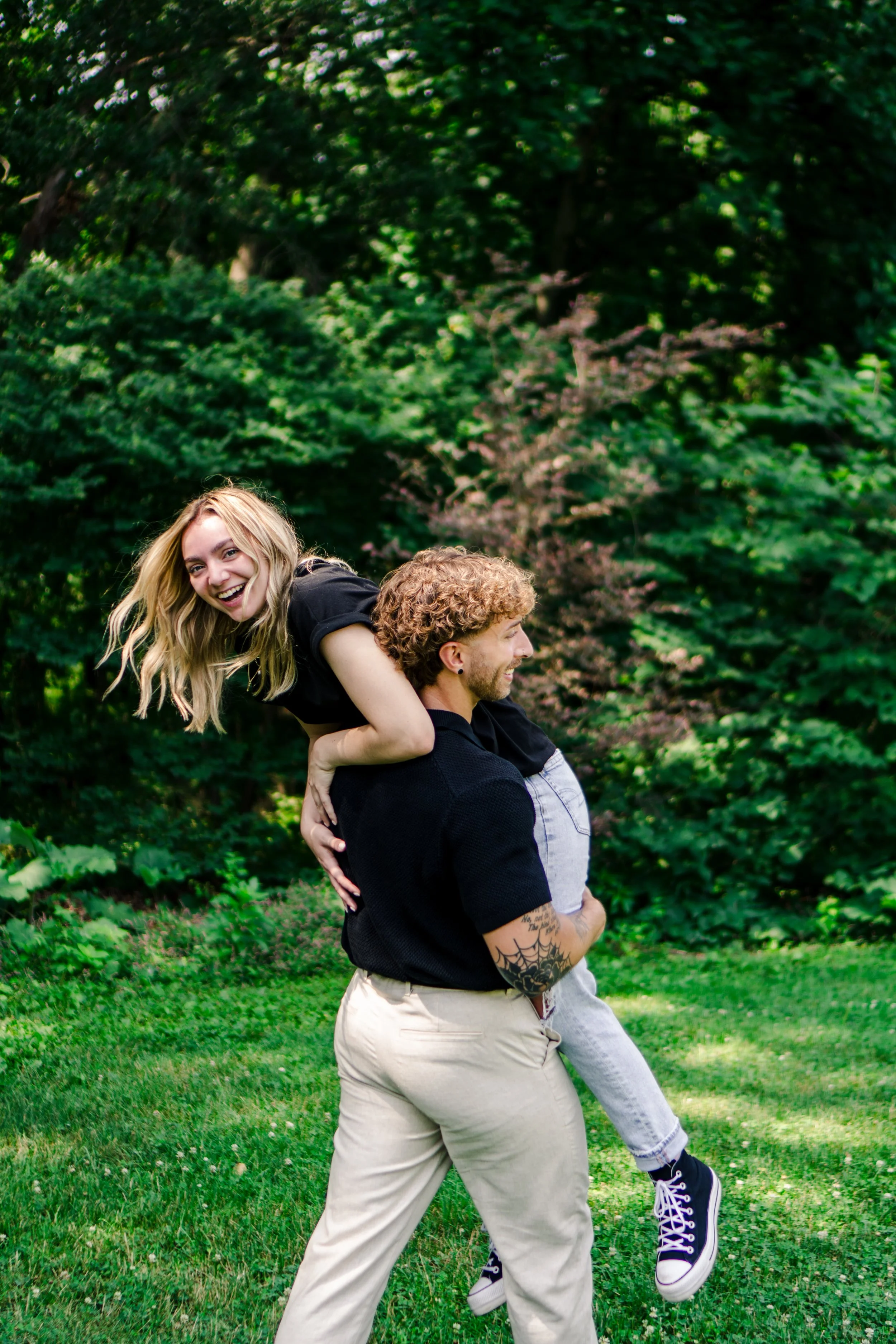 A man giving a piggyback ride to a woman in a park with lush green trees in the background. The woman is smiling and laughing, wearing a black shirt, while the man has tattoos on his left arm and is wearing a black shirt and beige pants.