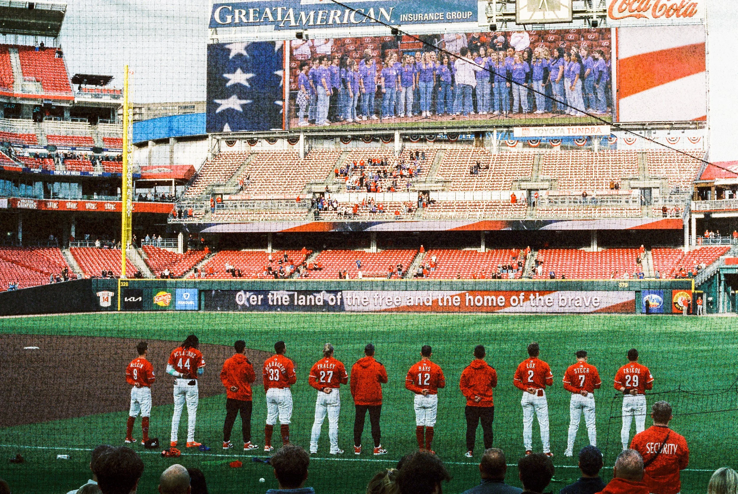 Baseball players in red uniforms standing on the field with audience in the stands. Large screen showing the team and American flag design, with banners and advertisements around the stadium.