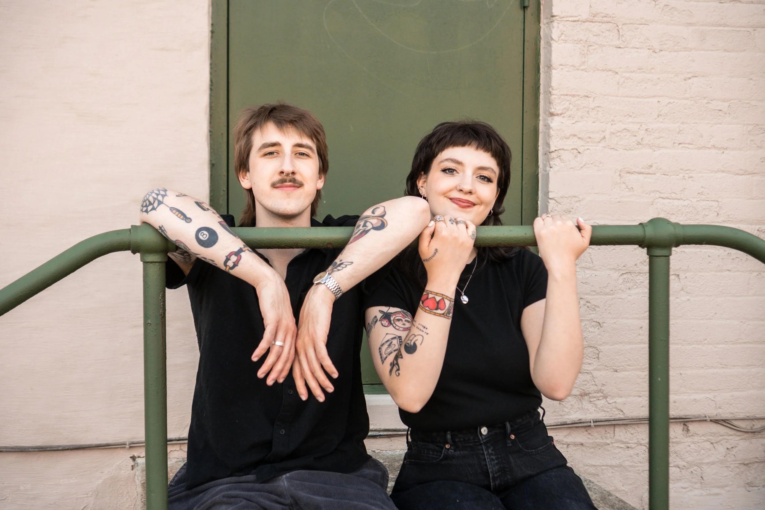 A young man and woman with tattoos leaning on a green railing in front of a brick wall and green door, smiling at the camera.