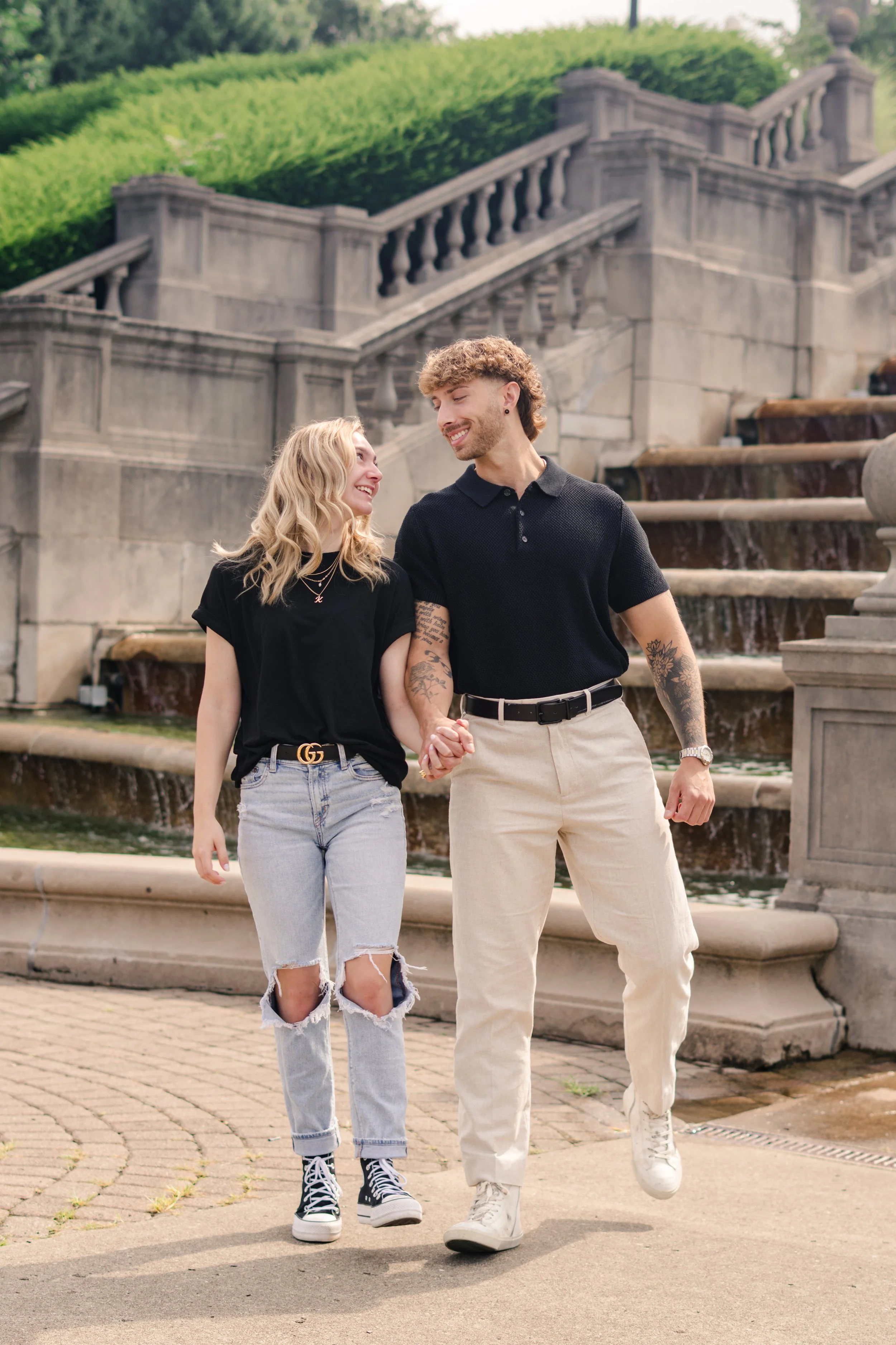 A young couple walking hand in hand outdoors near a stone fountain, both smiling and looking at each other. The woman has blonde hair, wears a black t-shirt, ripped jeans, and sneakers. The man has curly hair, tattoos, and is dressed in a black polo,