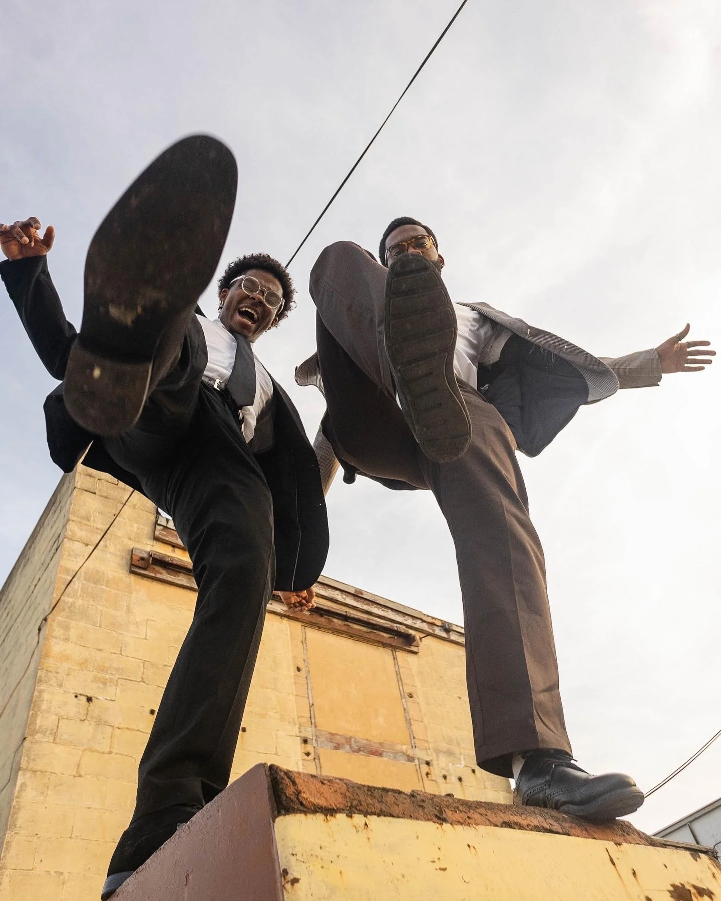 Two men in suits jumping and kicking towards the camera, standing on a rooftop with a yellow brick building behind them.