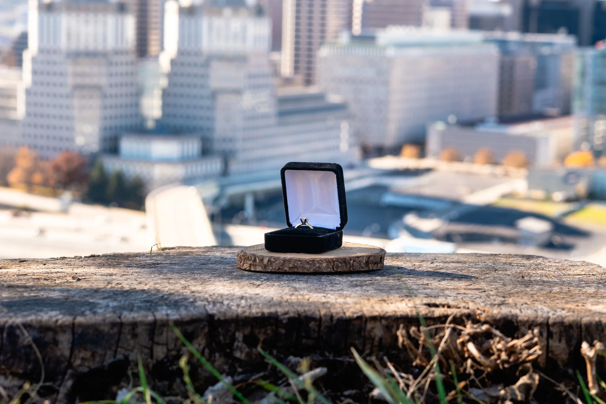 A black velvet jewelry box with a ring inside, placed on a piece of wood outdoors with city buildings in the background.