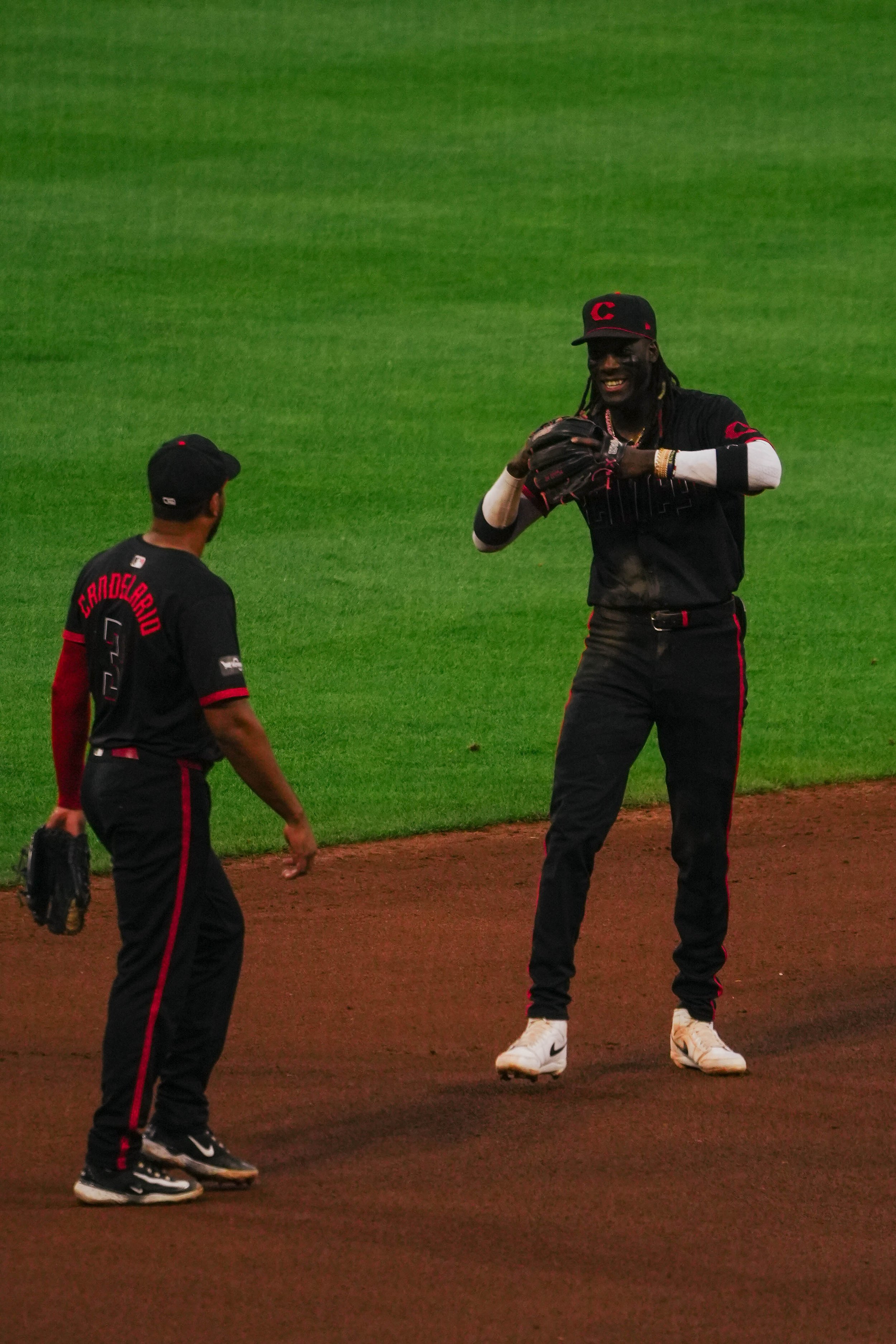 Two baseball players on the field, one is tall with long braids and a dark complexion, holding a glove and smiling, while the other, shorter, faces him with a glove in his left hand, both wearing black uniforms with red accents.