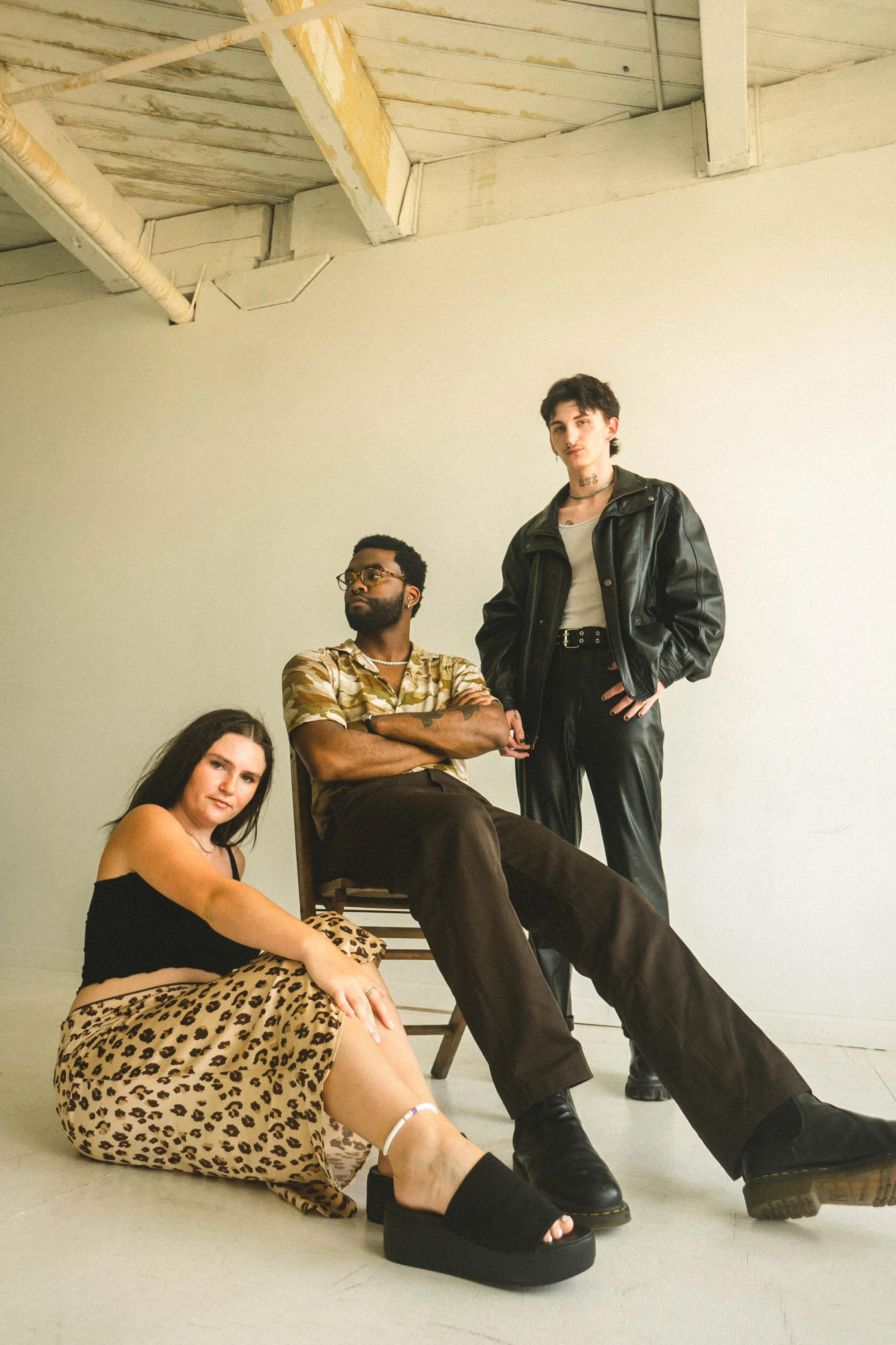 Three young adults posing in an indoor studio with a plain white wall and exposed ceiling beams, dressed in casual fashion.