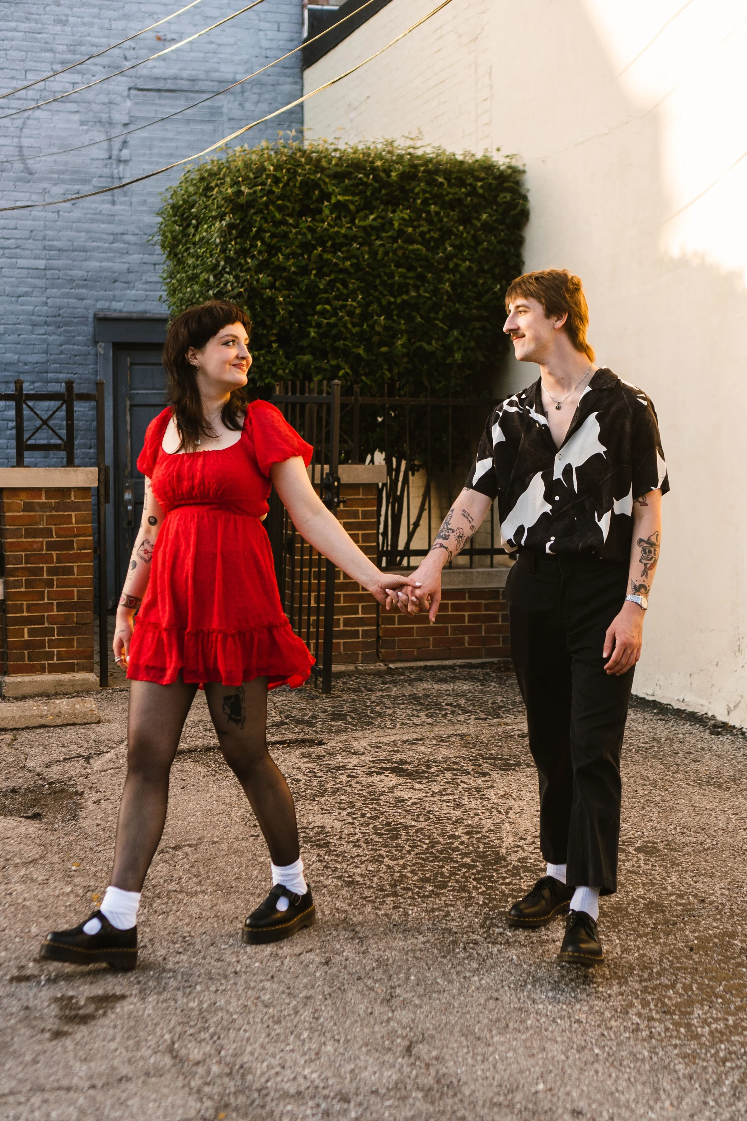 A young couple holding hands and smiling at each other in an outdoor alleyway at sunset. The woman is wearing a red dress, patterned tights, and black shoes, while the man is dressed in a black and white patterned shirt, black pants, and black shoes.