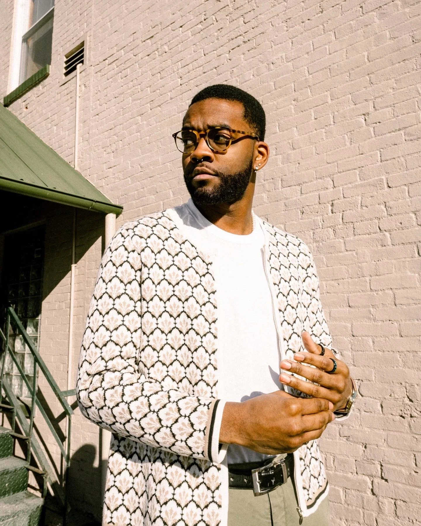 A man with glasses and a beard stands outdoors against a light brick wall, wearing a patterned jacket over a white shirt and holding his hands together.