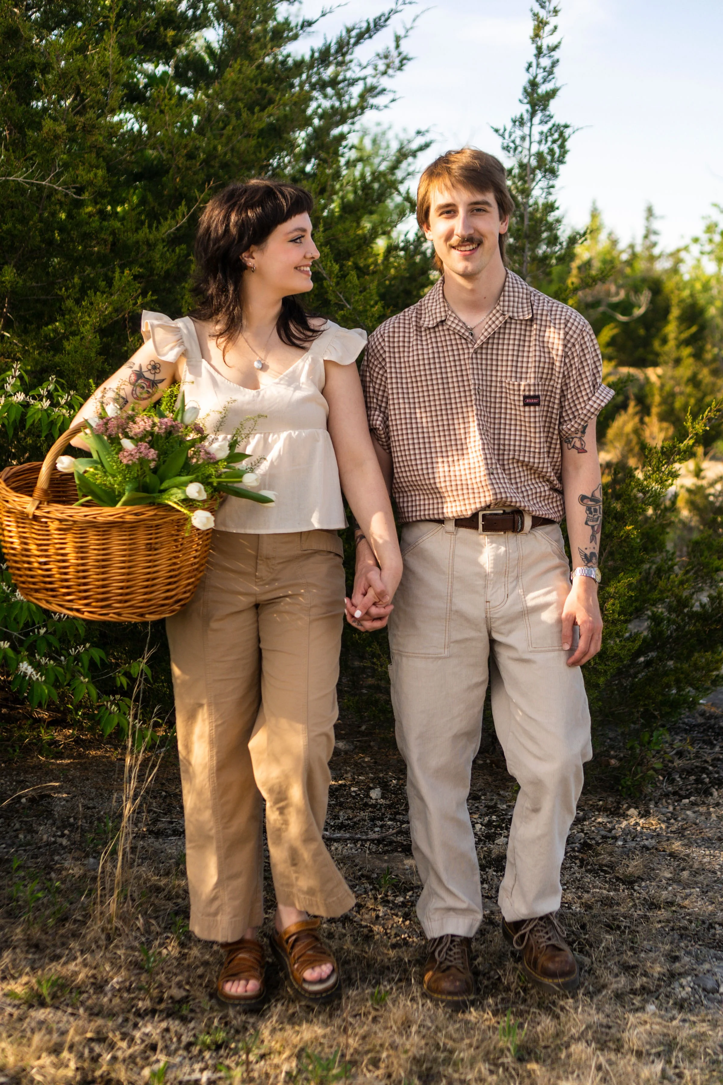 A young couple walking outdoors holding hands, with the woman carrying a basket of flowers and greenery, surrounded by lush green trees, on a sunny day.