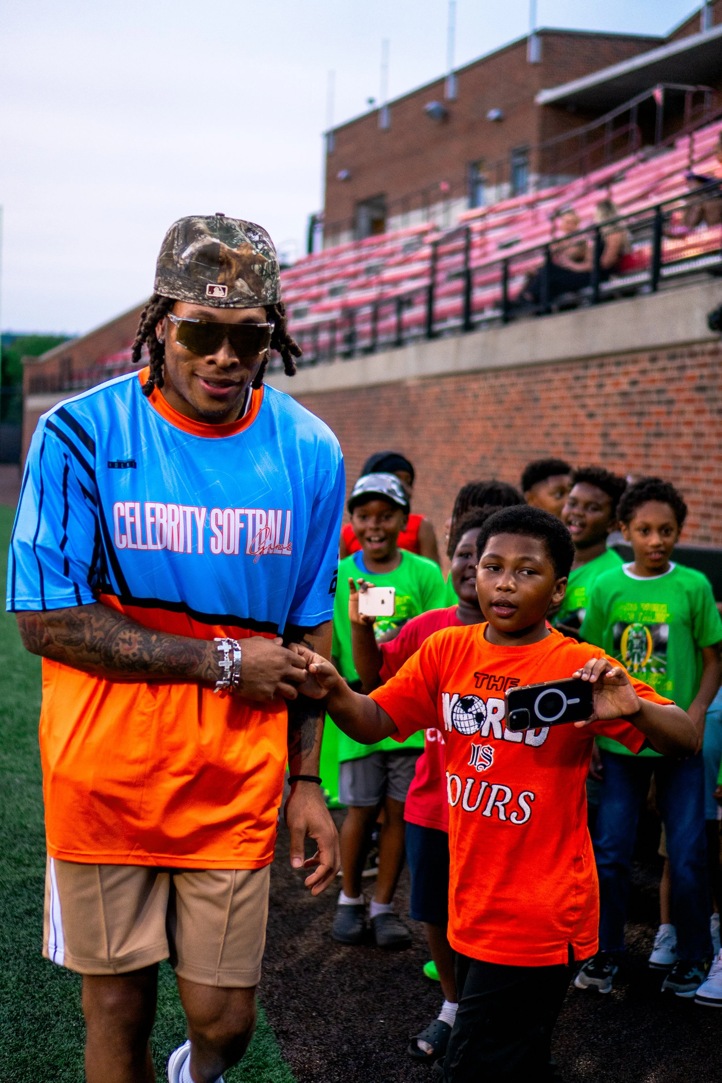 A man with sunglasses and tattoos on his arm is shaking hands with a young boy in an orange shirt, surrounded by children smiling and holding phones on a sports field.