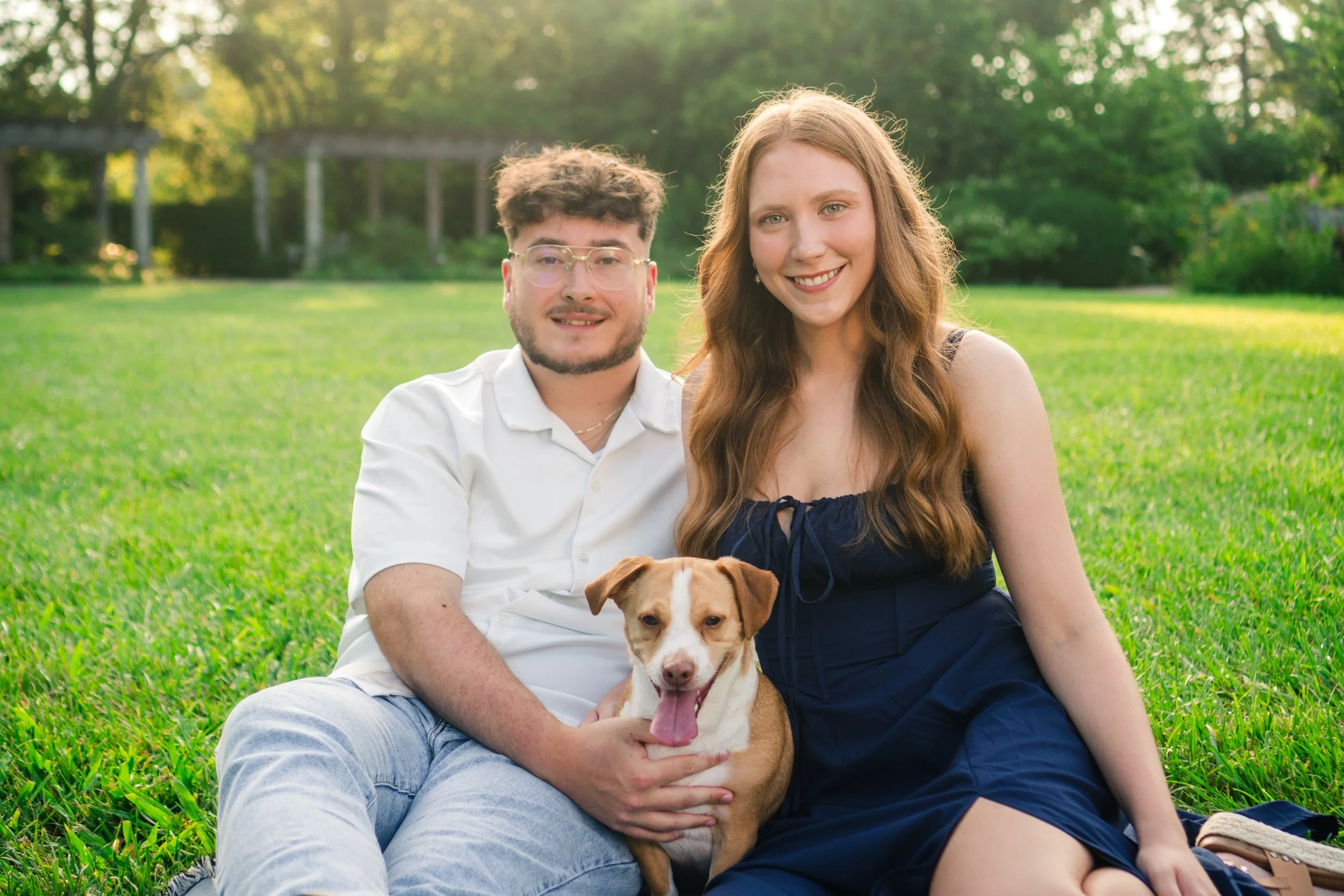 A happy couple sitting on the grass in a park with their dog, surrounded by trees and greenery.