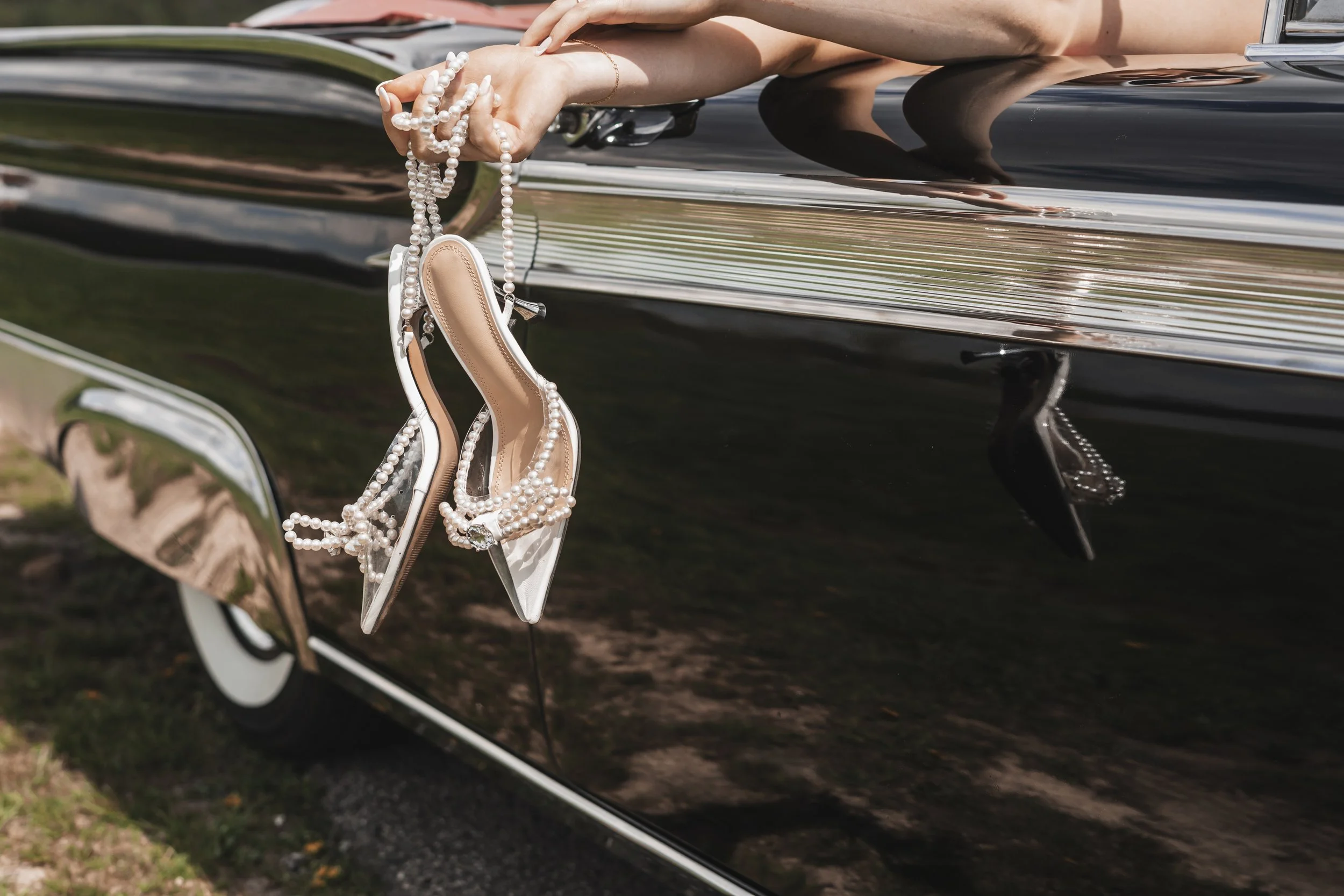 A pair of white high-heeled shoes decorated with pearl necklaces hanging on the side of a shiny black car.
